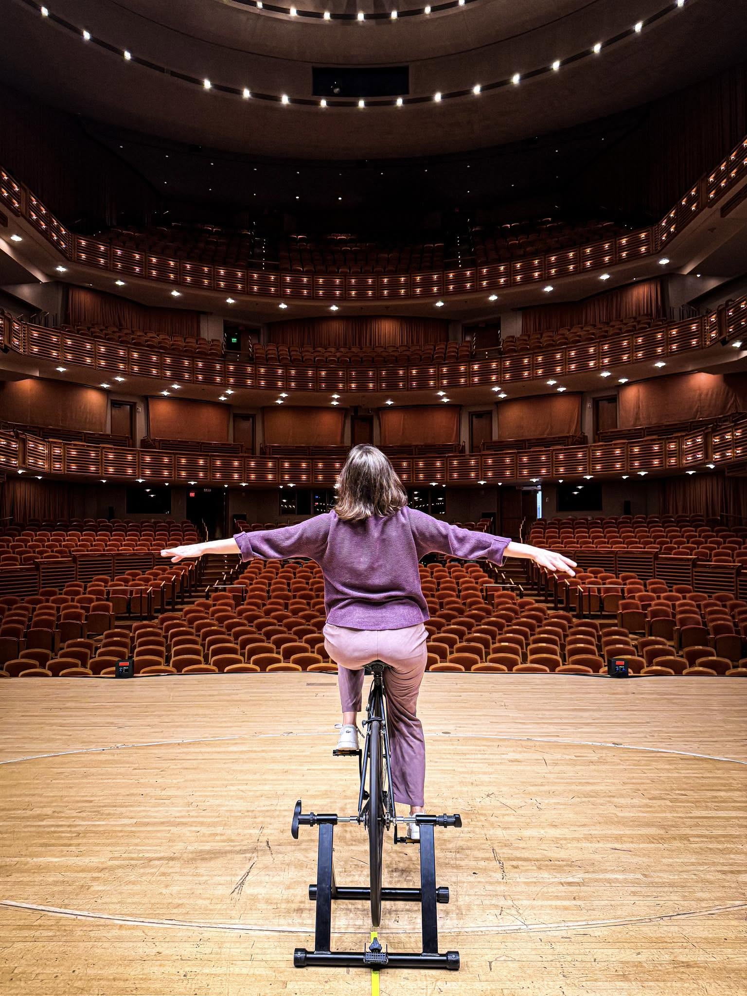 Child balancing on a bicycle on stage in an empty theater with multiple balconies and rows of seats.