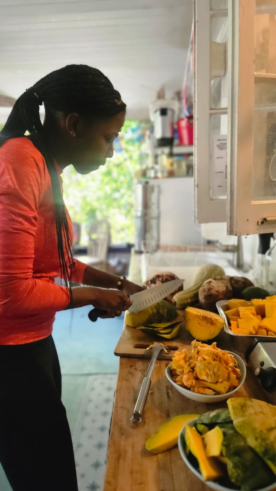 Person with dark hair in a ponytail, wearing an orange top, preparing food in a kitchen with various vegetables and a cutting board.