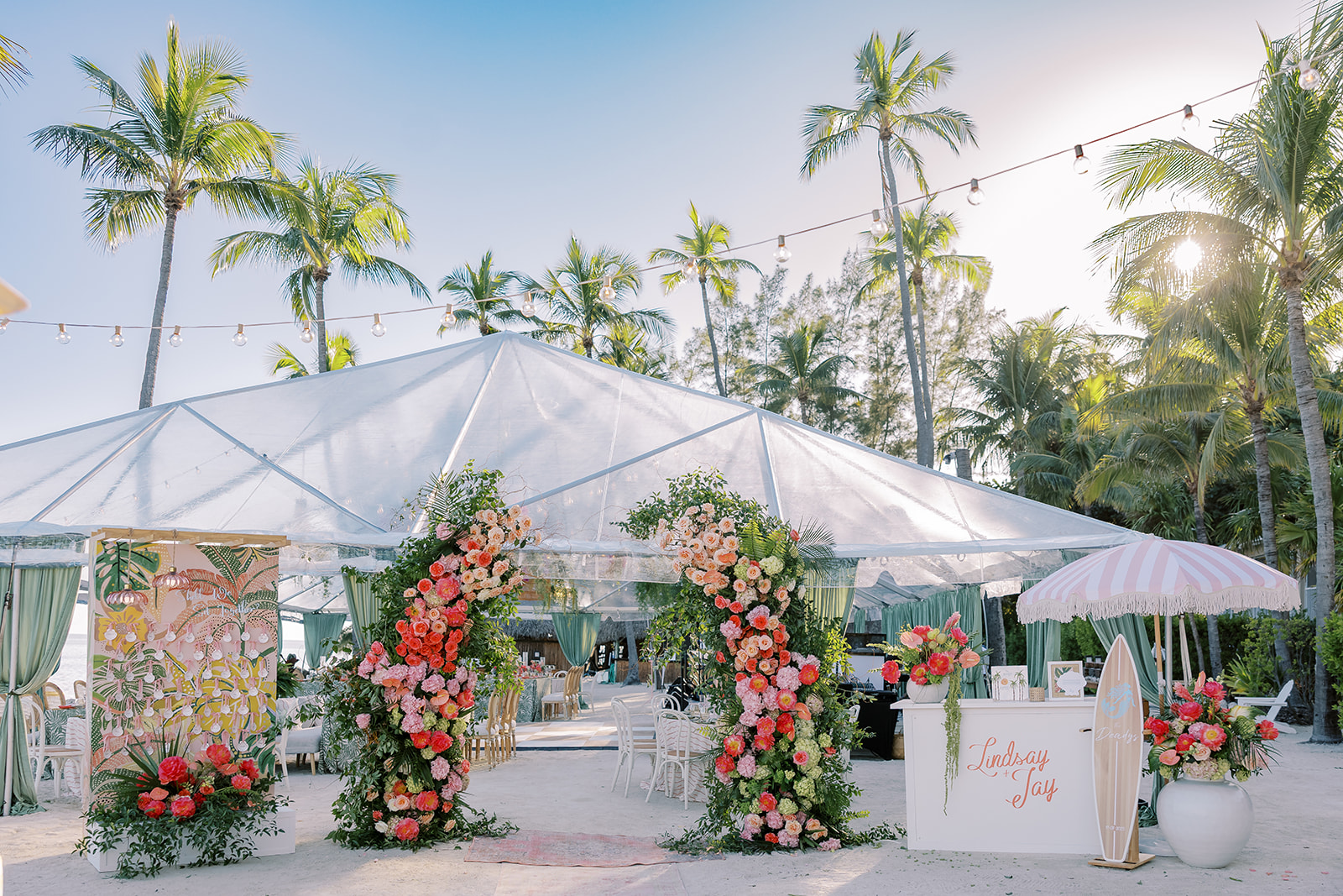 Outdoor event space with palm trees, decorated with pink and white flowers, under a clear sky.