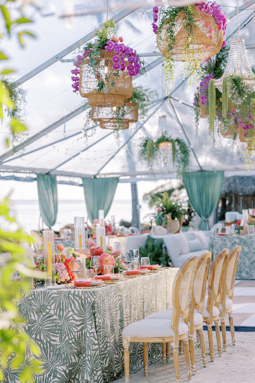 Elegant indoor event with decorated table, hanging floral arrangements, and draped curtains, under a glass ceiling.