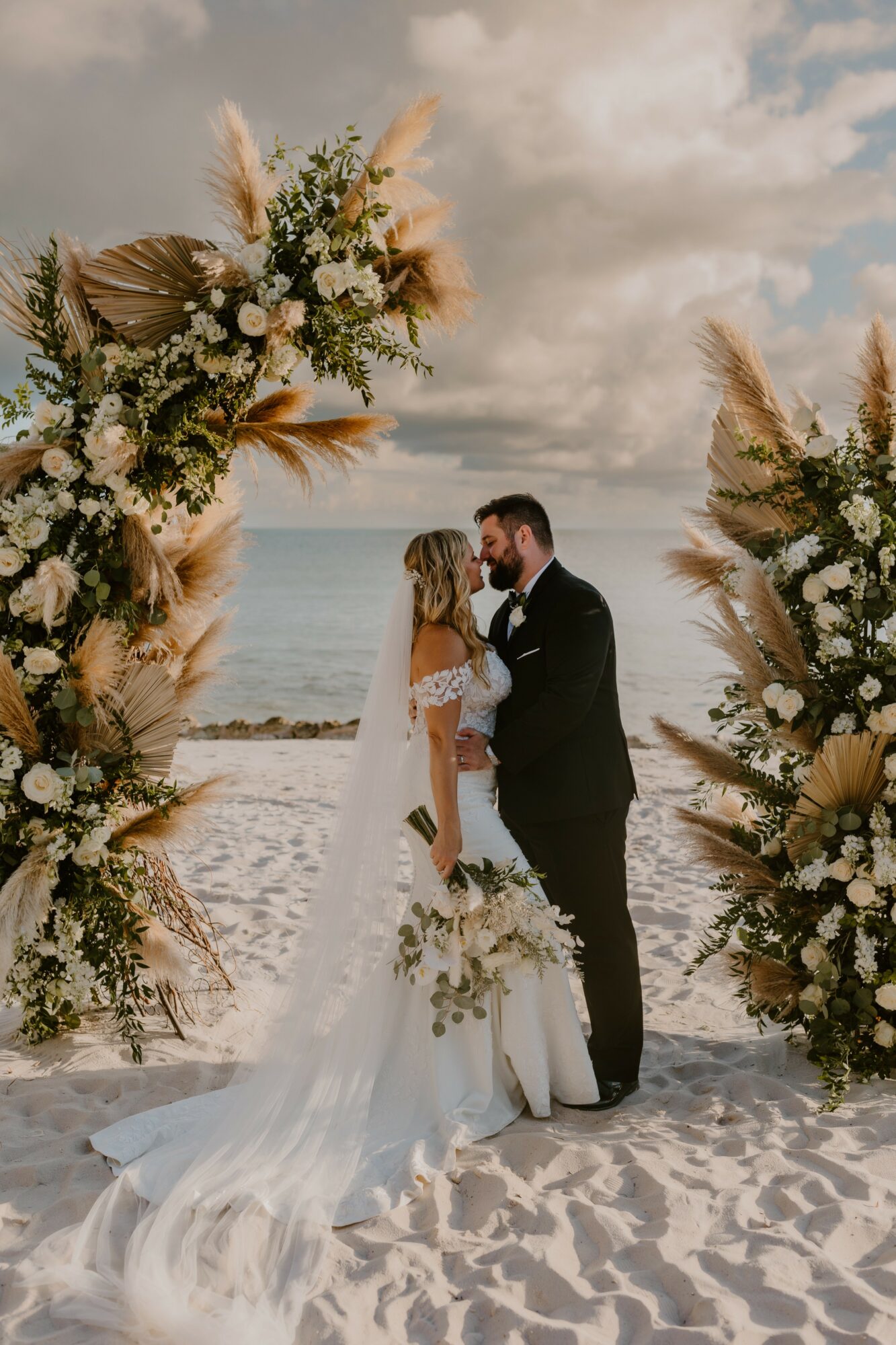 Bride and groom standing on a beach, holding hands, surrounded by large floral arrangements, with ocean and cloudy sky in background.