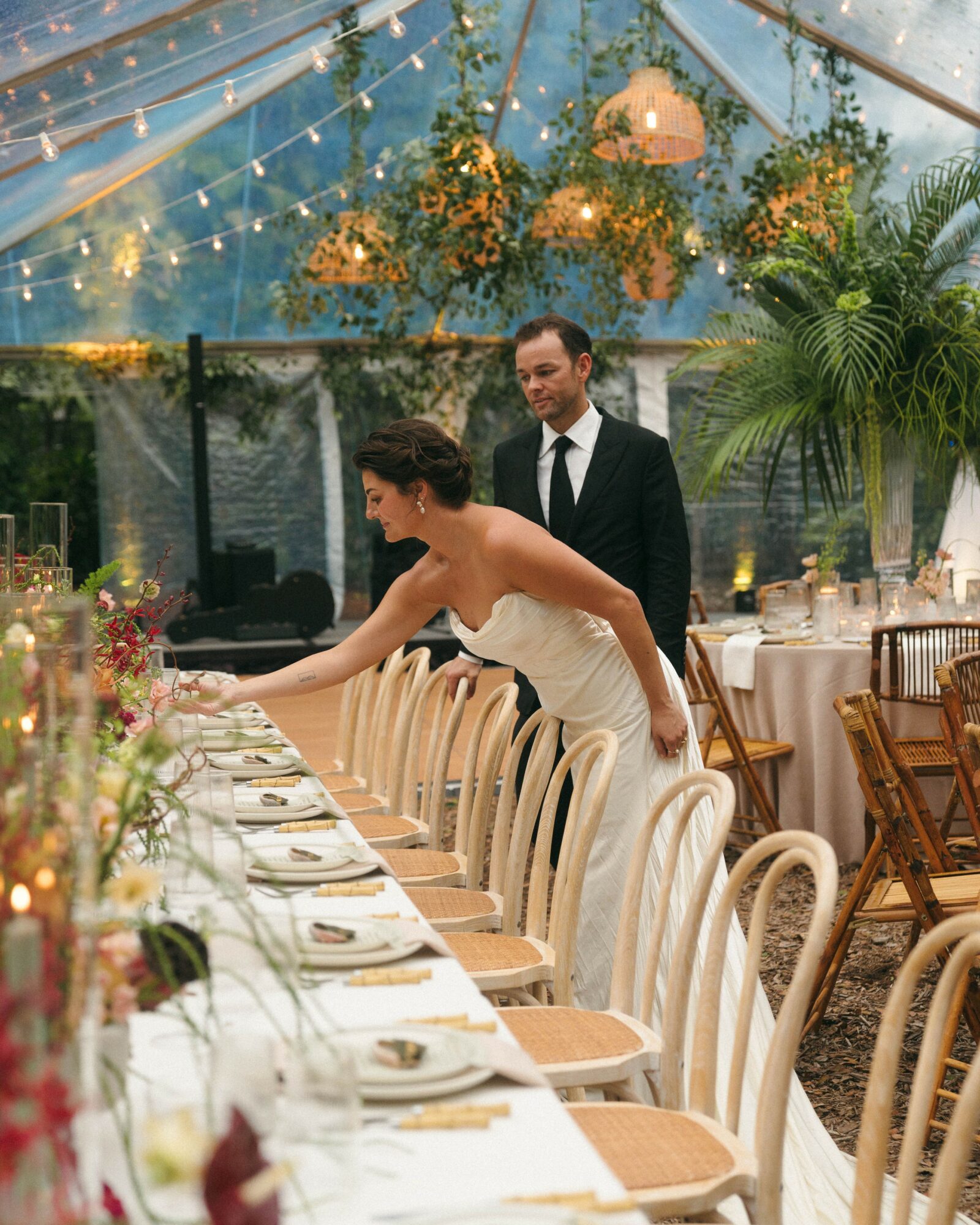 Woman in wedding dress reaching for table setting, man in suit standing behind her, decorated outdoor event space with plants and lights.