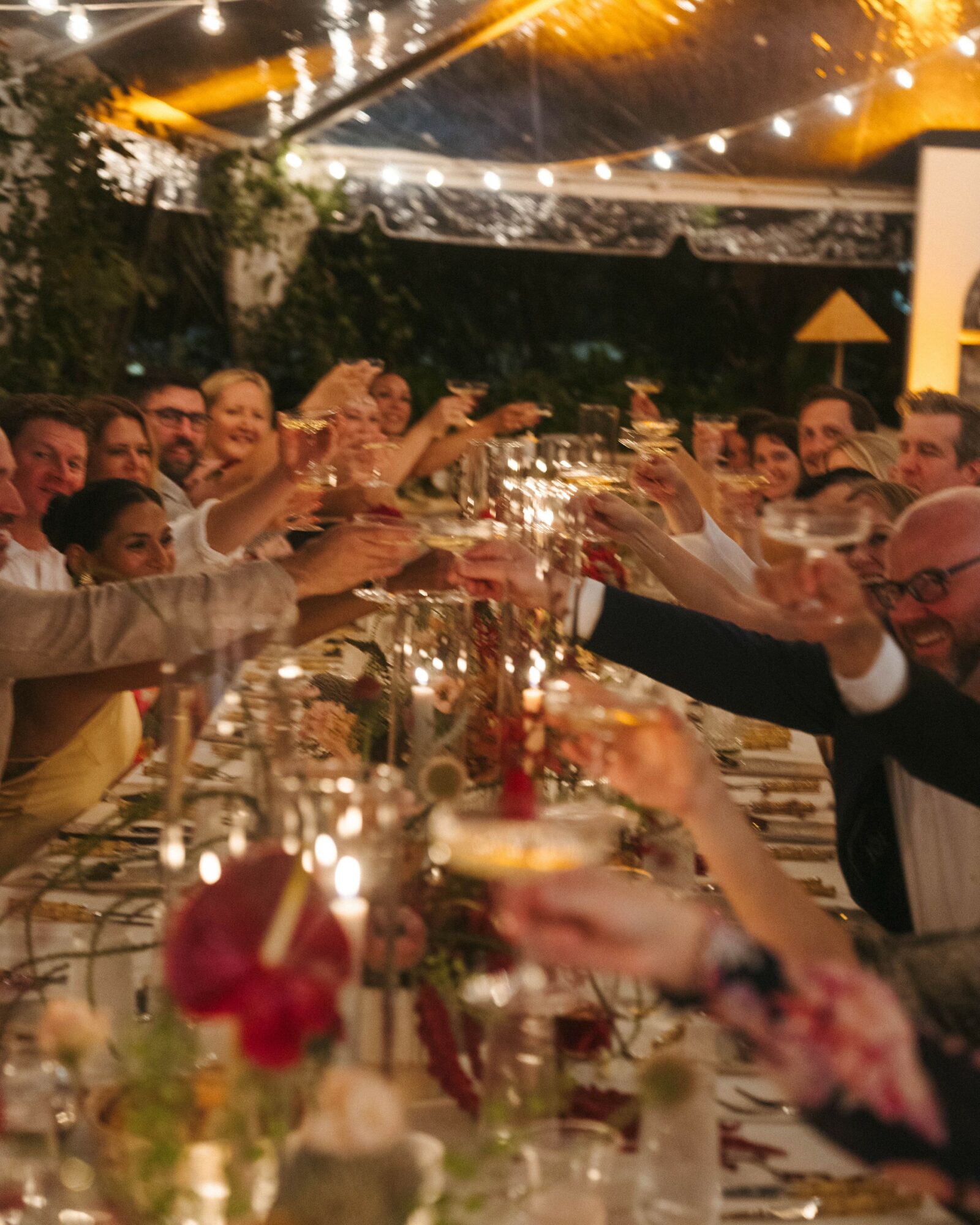 People raising glasses in a toast at a festive outdoor dinner party with string lights overhead.