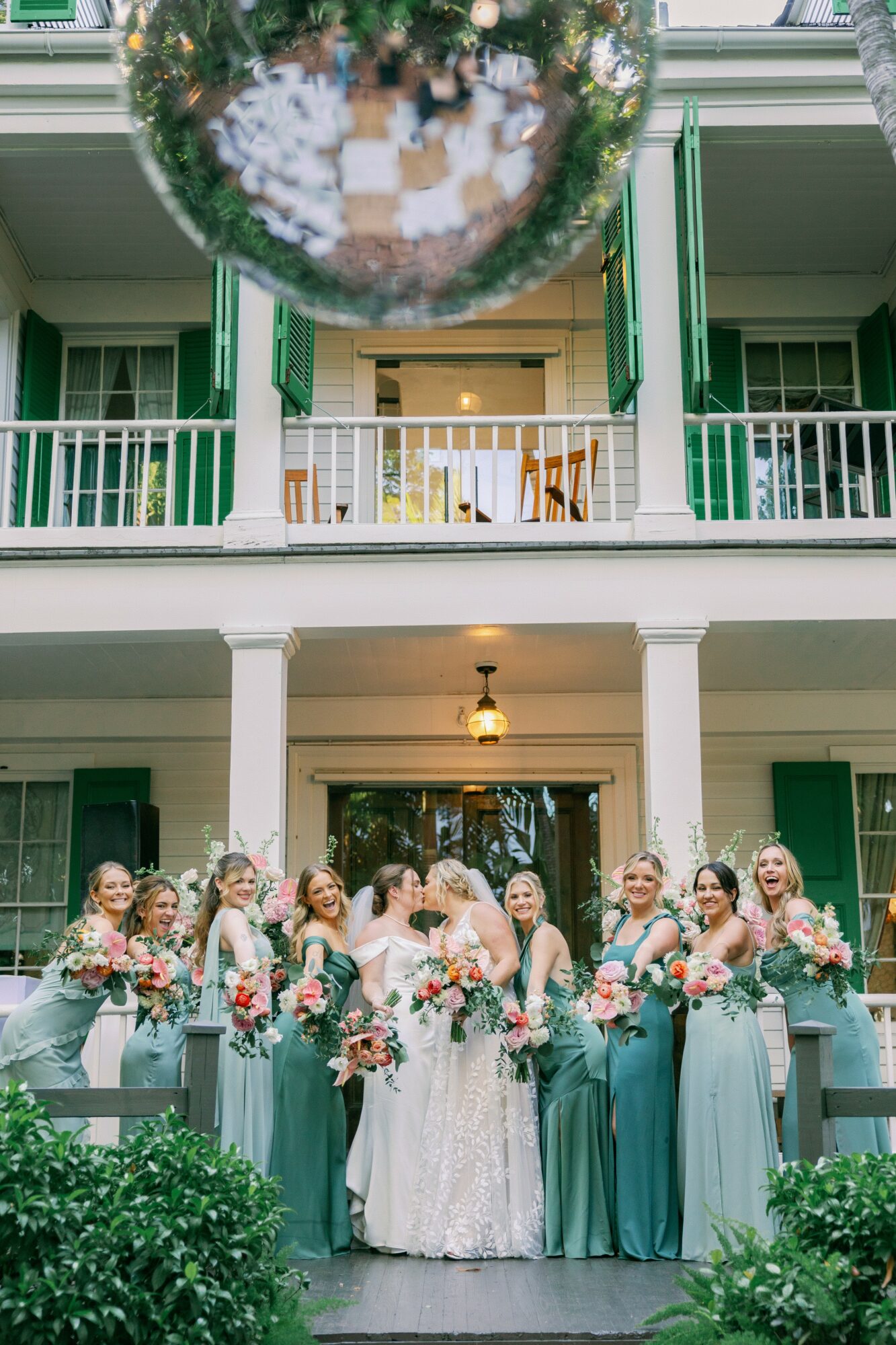 Group of women in formal dresses and a bride in a white gown holding bouquets, standing outside a building with green shutters.