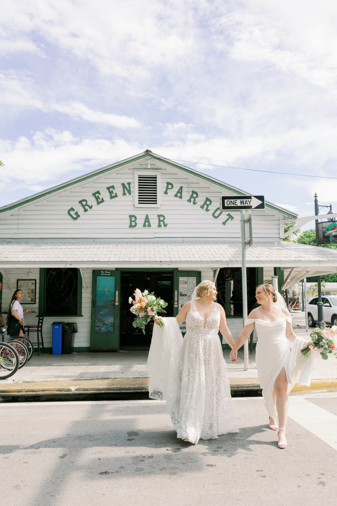 Three women in white dresses walk across a street in front of a building with a sign reading 'Green Parrot Bar.'