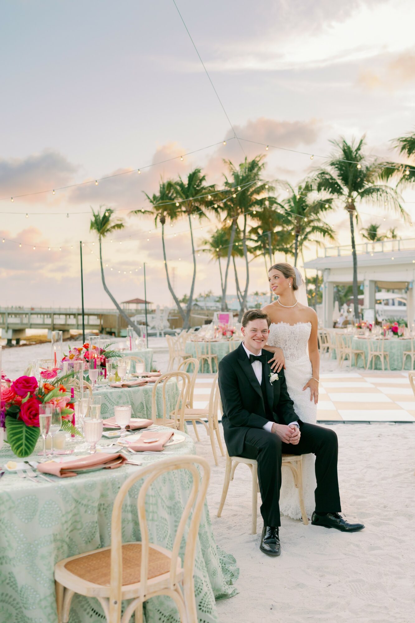A bride and groom pose outdoors near decorated tables with flowers, palm trees, and a marina in the background during sunset.