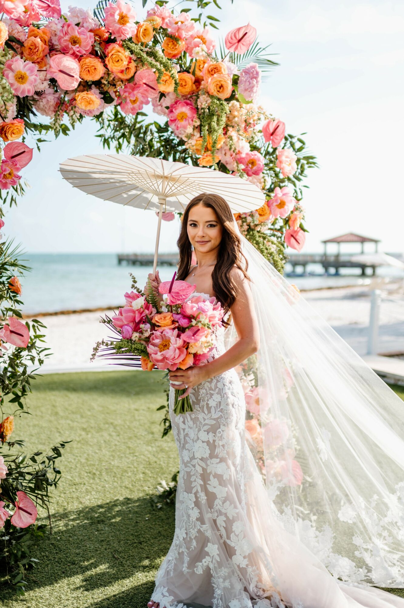 Bride in a lace wedding dress holding a bouquet, standing outdoors near water with a floral arch and umbrella, sunny day.