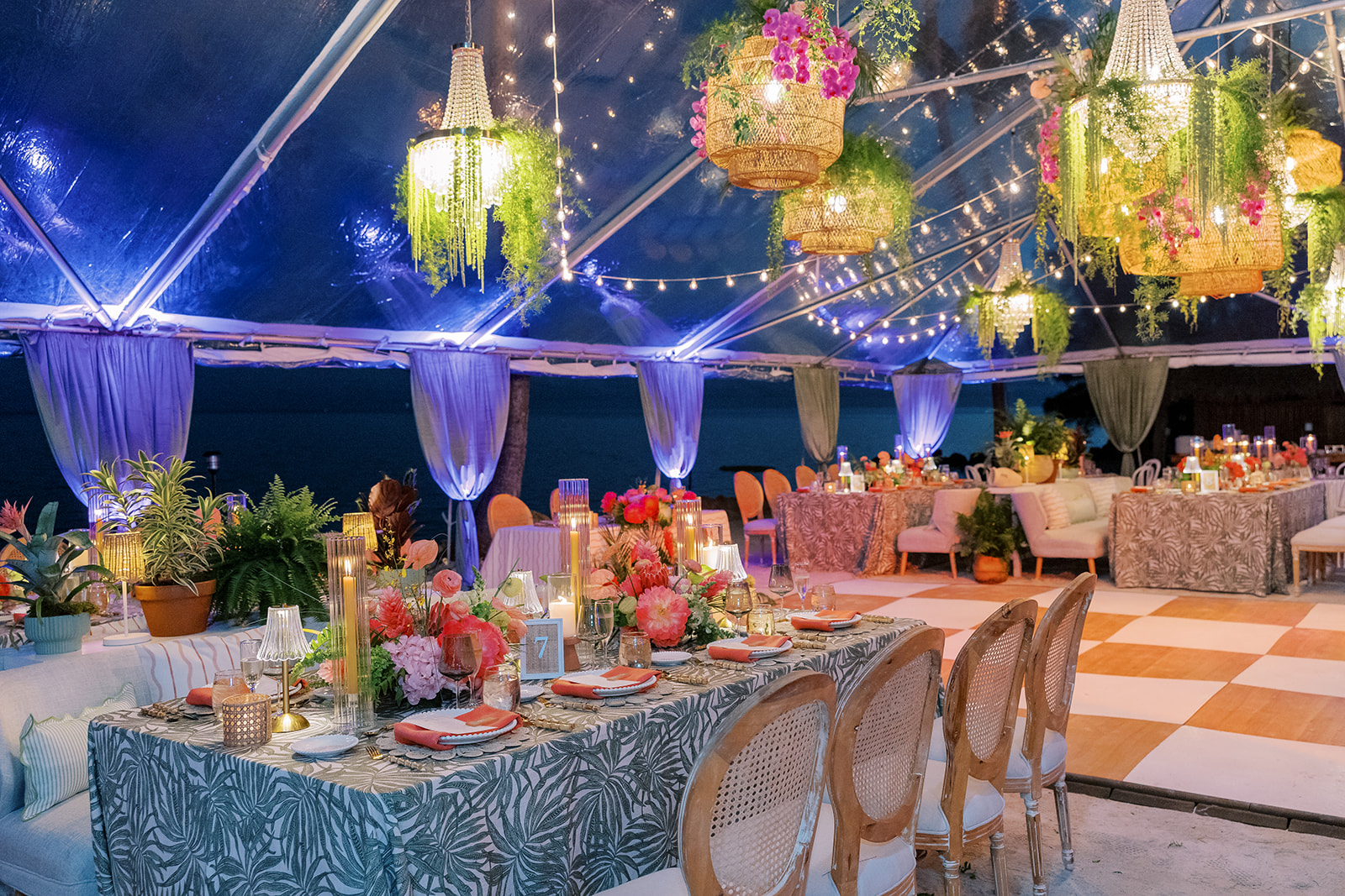 Decorated outdoor event space with hanging floral arrangements, string lights, and tables with floral centerpieces and candles.