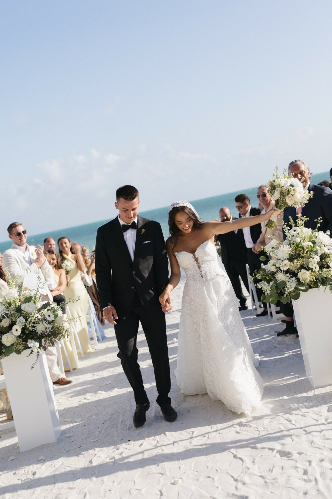 Bride and groom holding hands on a beach, surrounded by guests and floral arrangements, with ocean and sky in background.