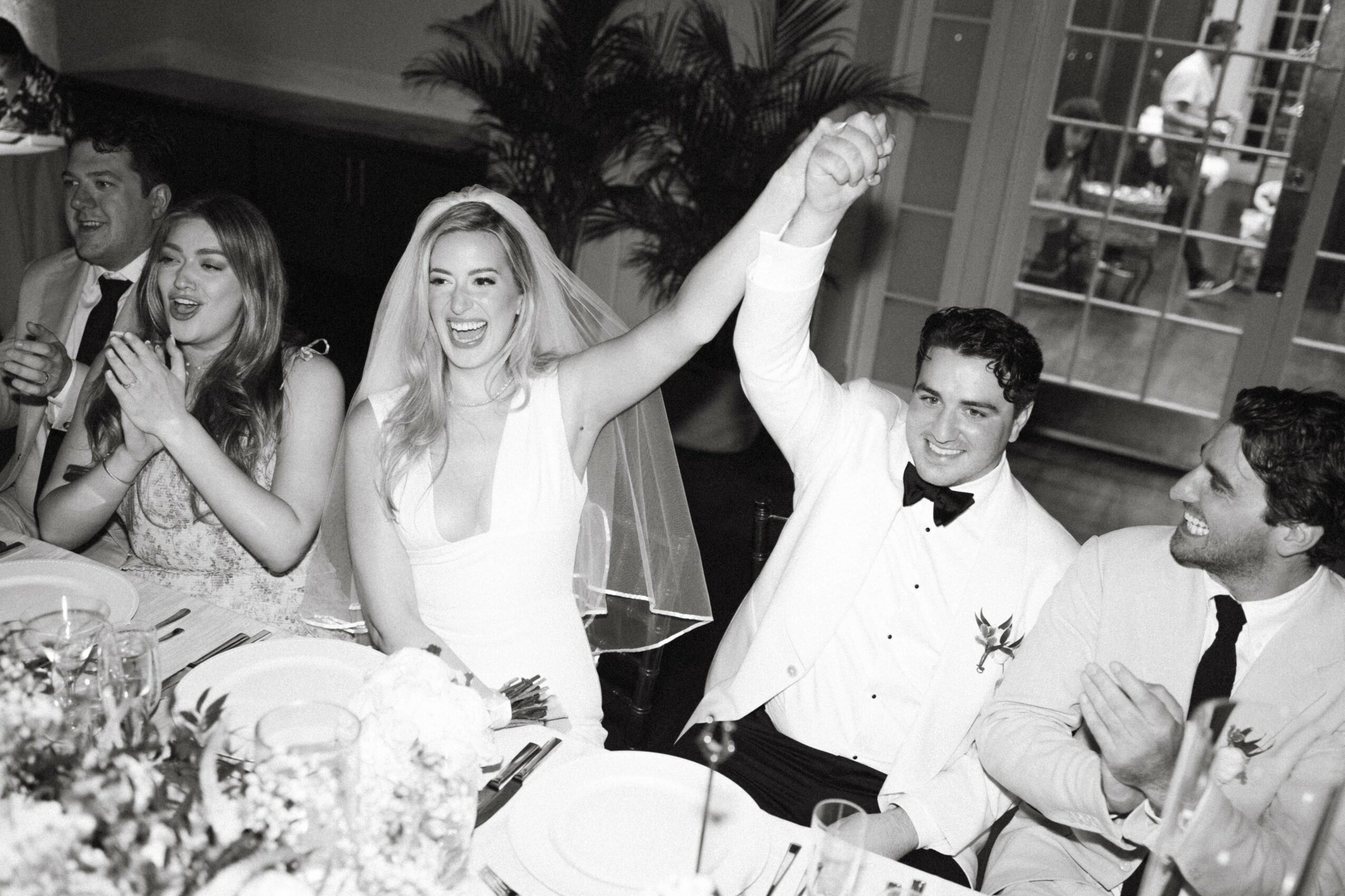 Group of people celebrating at a wedding reception, with a bride and groom raising their hands, smiling, and clapping.