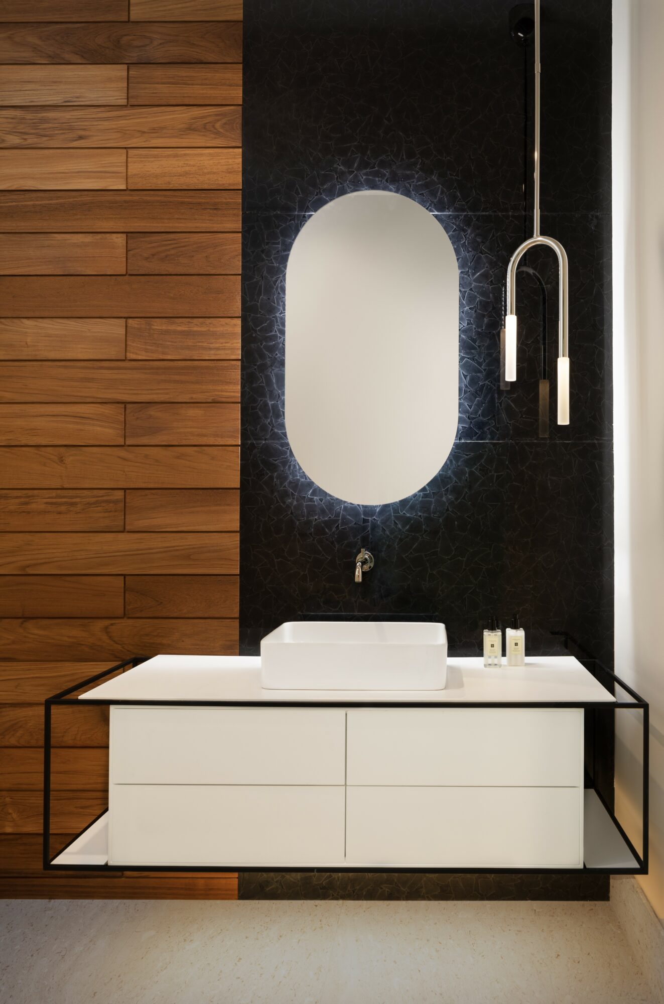 Bathroom vanity with a white sink, oval mirror, black textured wall, wooden panel, and modern hanging faucet.