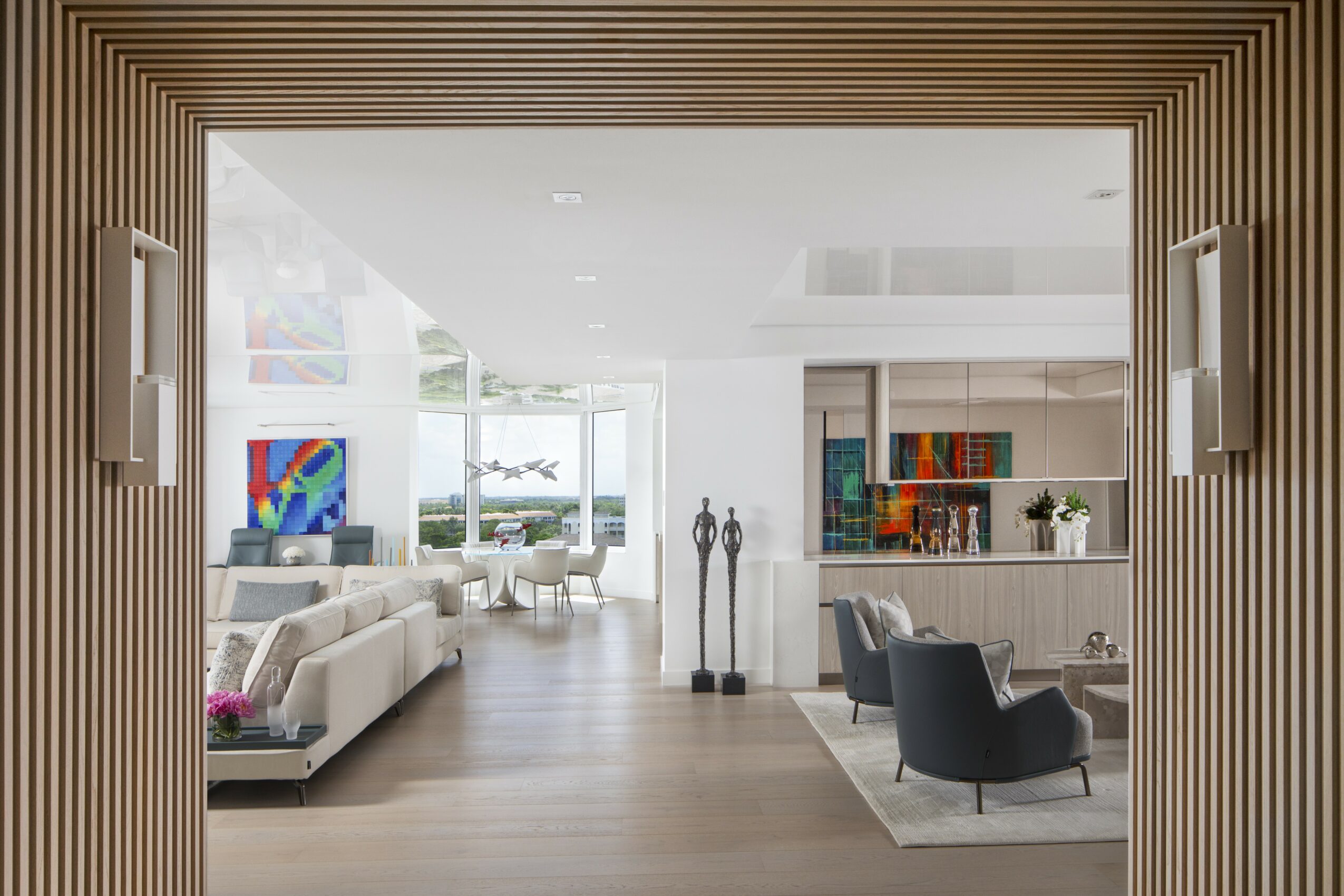 Living room with white sofa, black armchair, artwork, and large windows, viewed through a wooden frame.