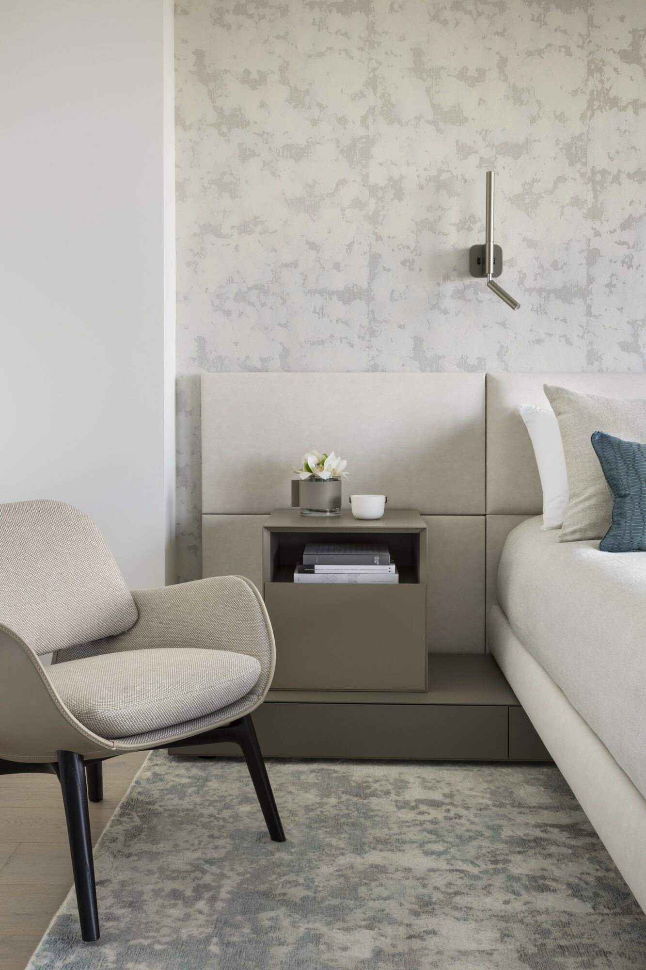 Bedroom with a bed, nightstand, and armchair, featuring neutral tones and textured wall, with a clock on the wall.