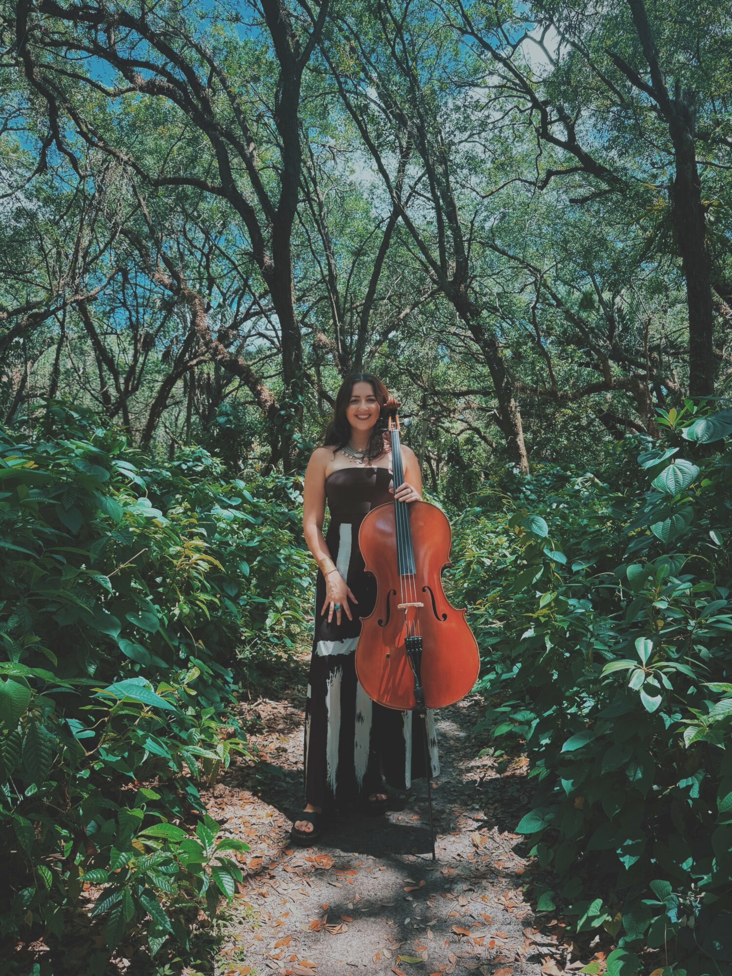 Woman standing on a forest path holding a cello, surrounded by lush green trees and foliage.