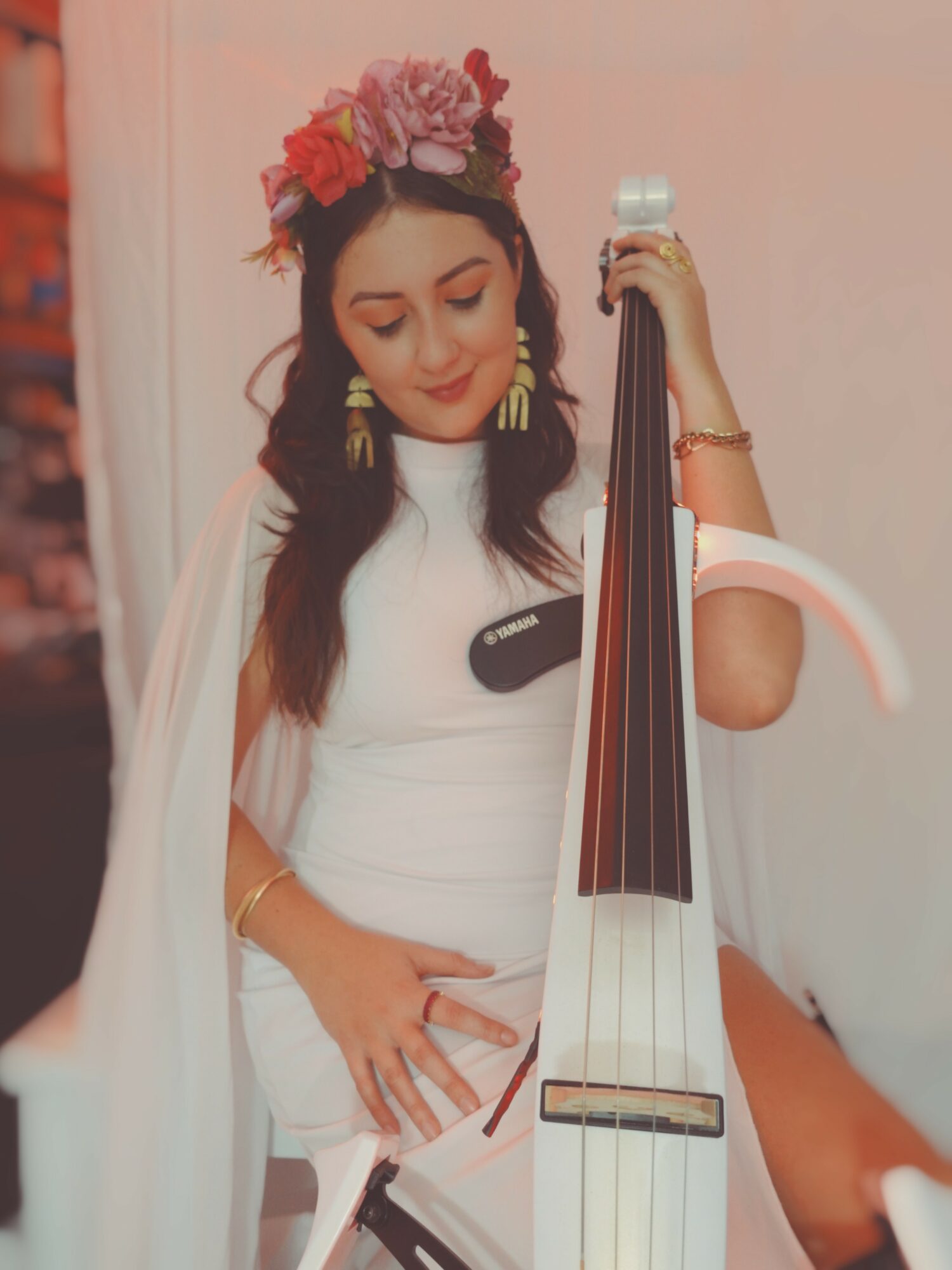 Woman with floral headband playing a string instrument, wearing a white dress, smiling, with a plain background.