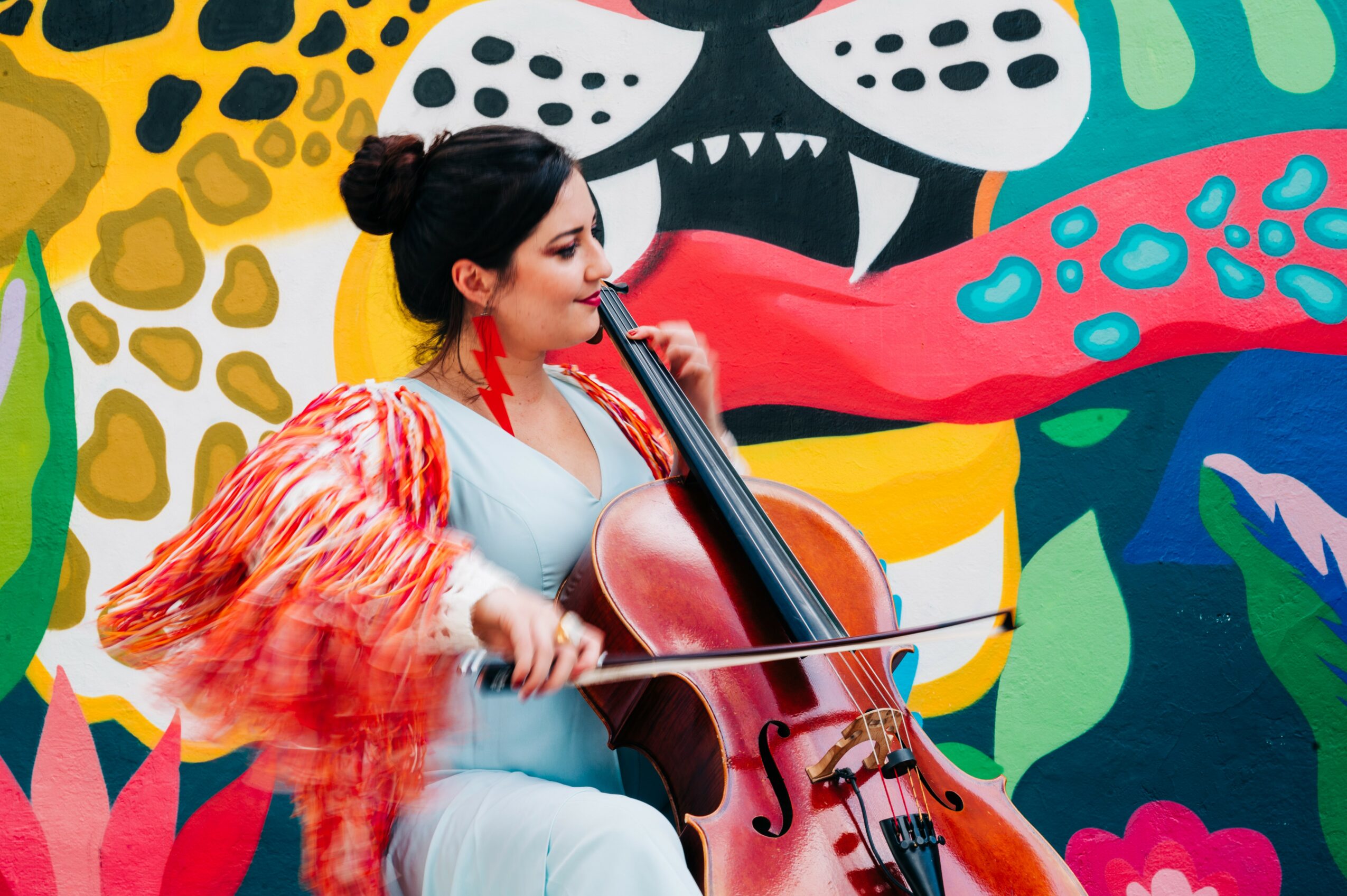Woman playing a cello in front of a colorful, abstract mural background.