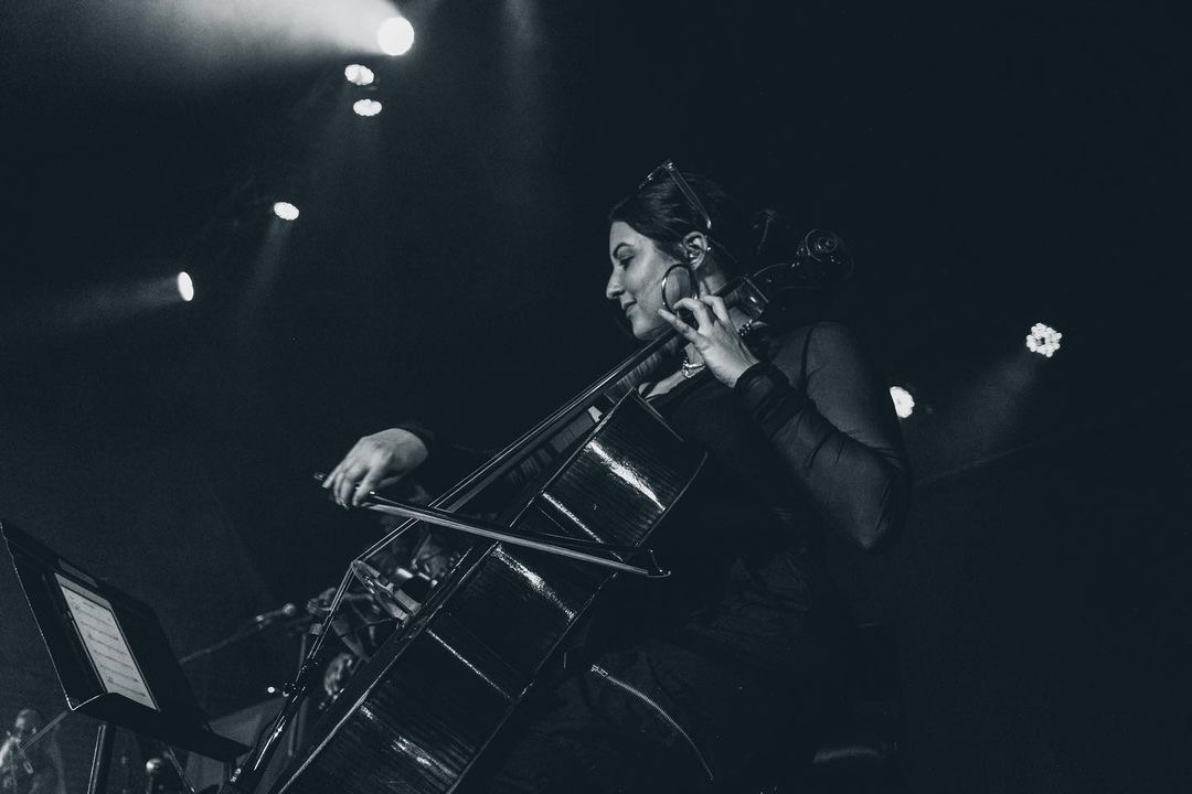 A woman plays a cello on stage with stage lights overhead, in black and white, focused on her performance.