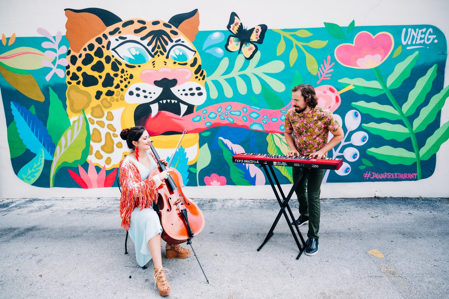 Two musicians perform in front of a colorful jungle mural with a leopard, butterfly, and pink flower. One plays a cello, the other a keyboard.