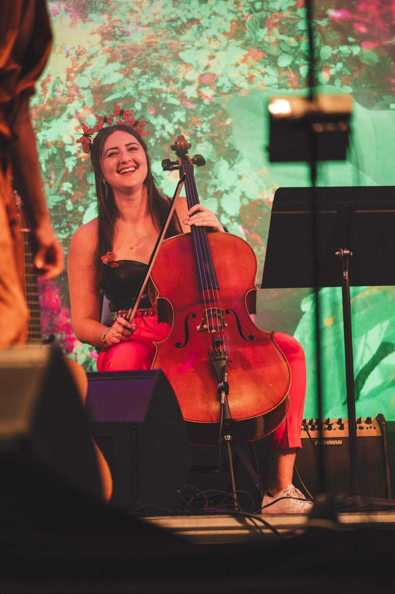 Woman smiling while holding a cello on stage with a colorful background, music stand, and microphone nearby.