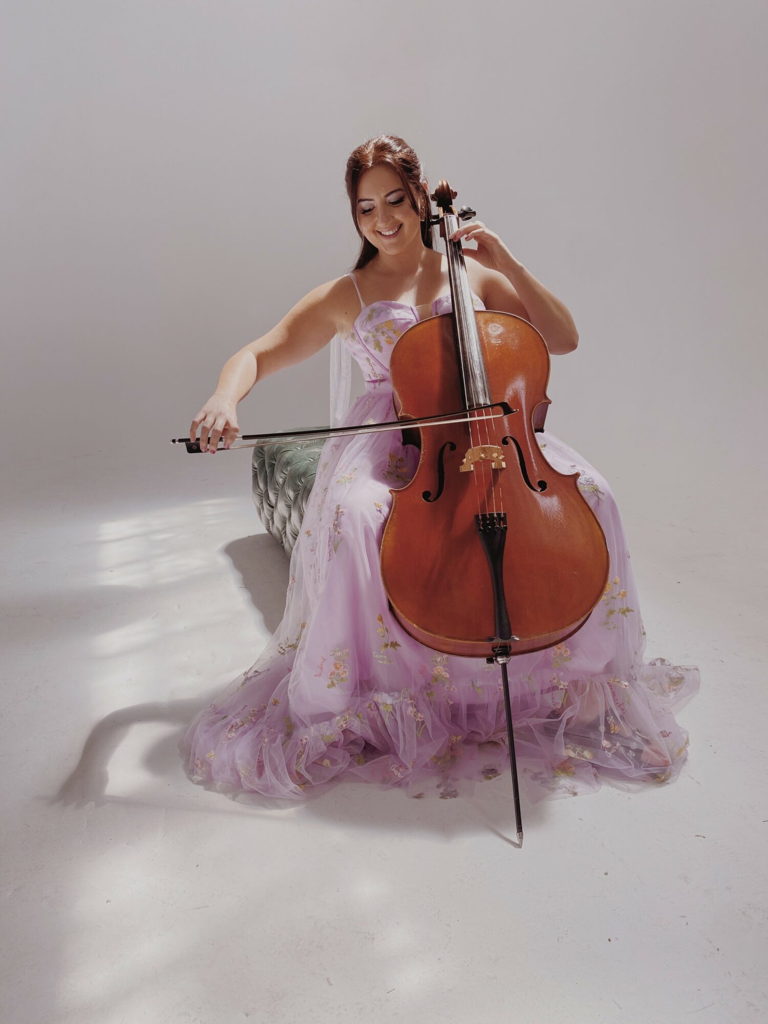 Woman in a pink dress playing a cello, sitting on the floor against a plain background.