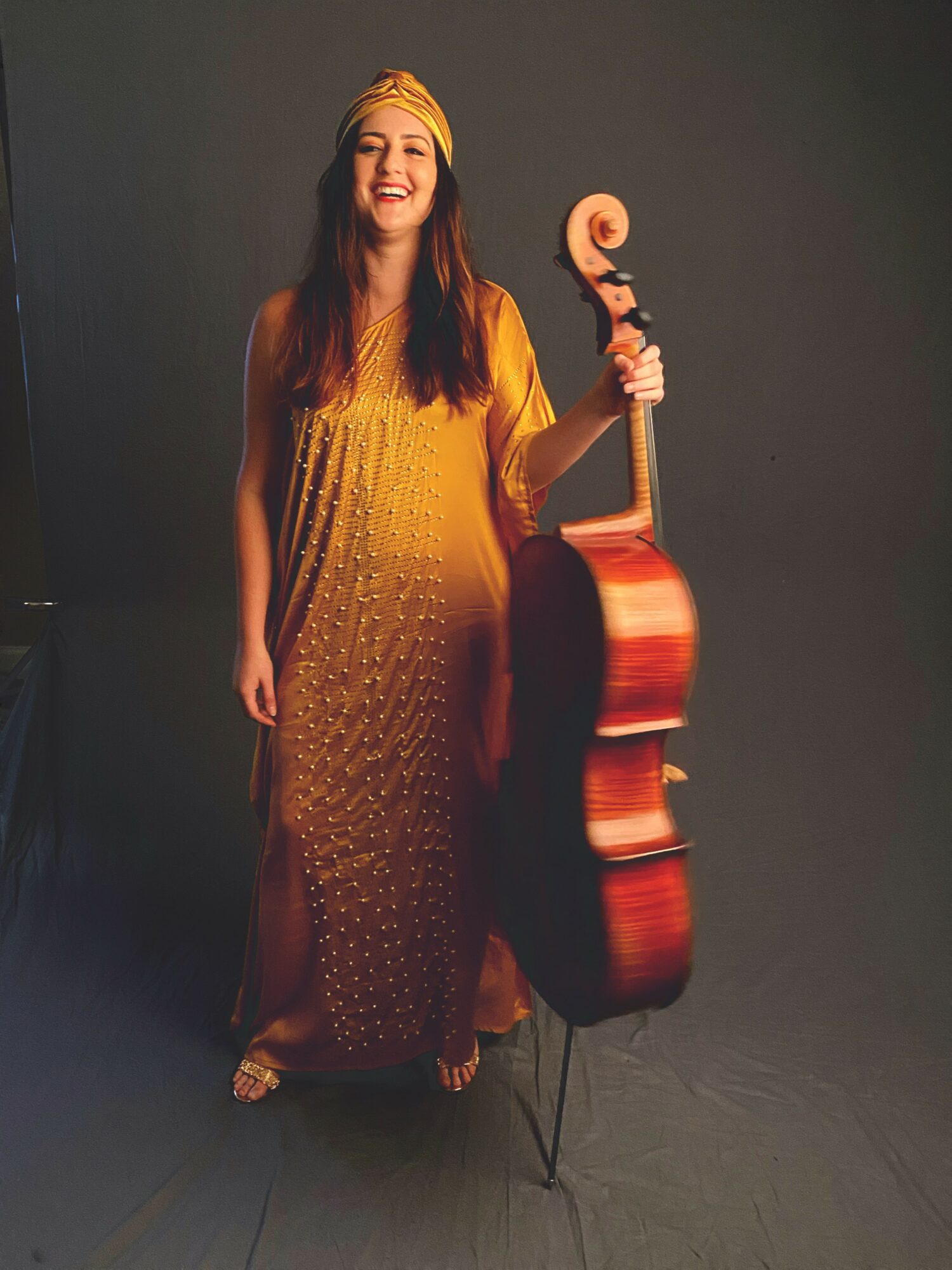 Woman in yellow dress and headband smiling, holding a large wooden acoustic guitar, standing against a dark background.