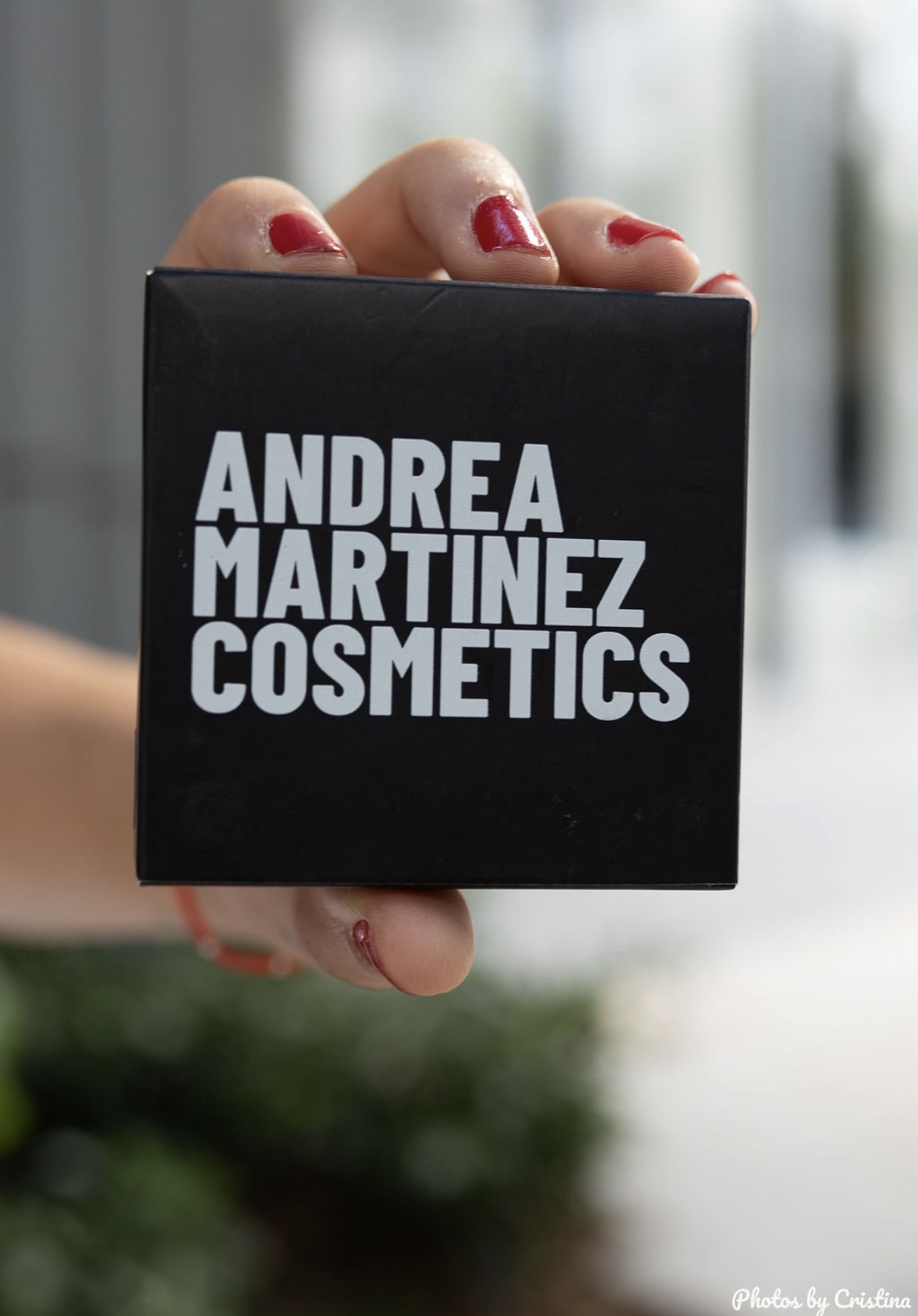 Person holding a black box with white text reading 'Andrea Martinez Cosmetics' in front of a blurred background.