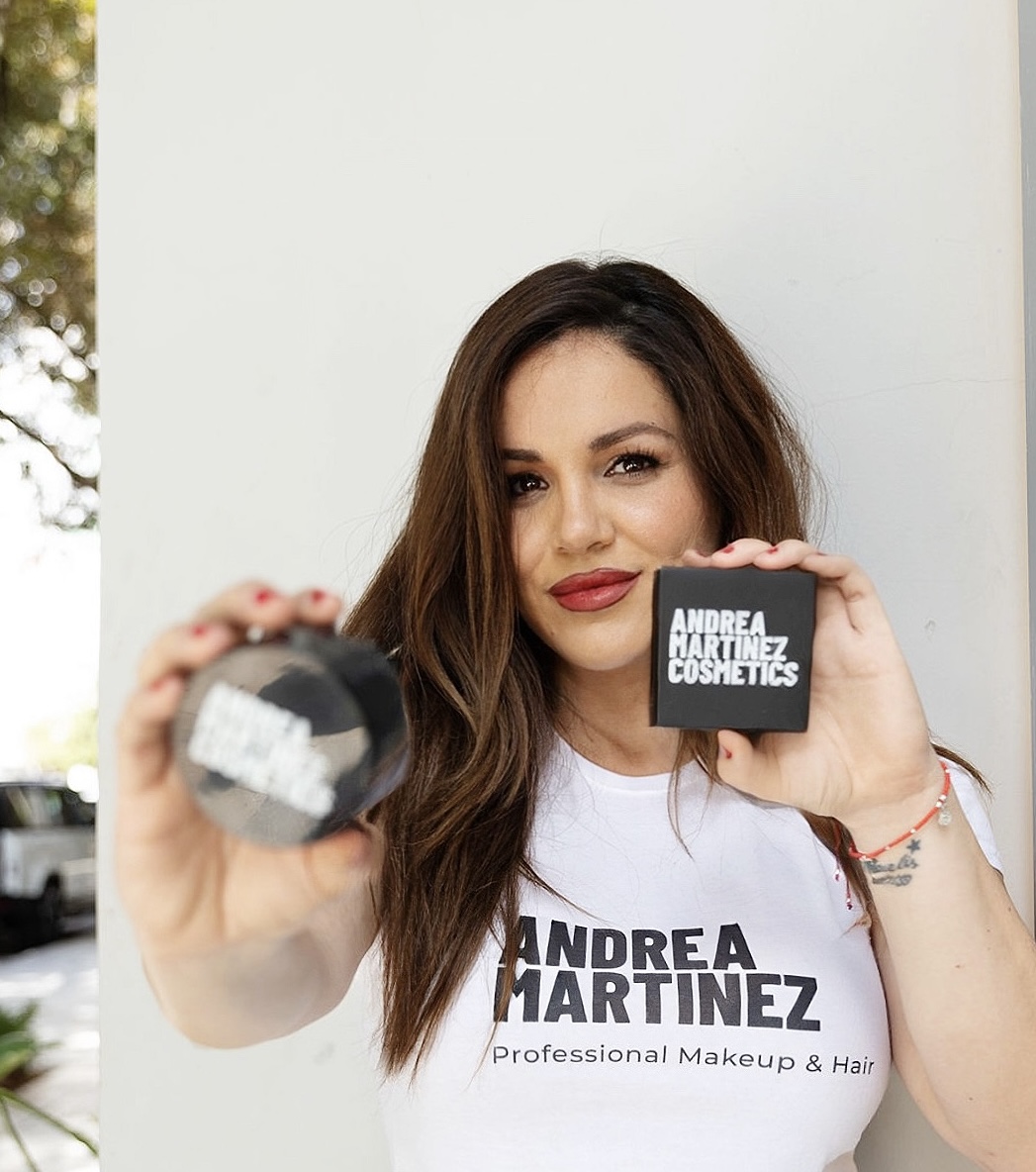 Woman holding a makeup product and a sign with her name, smiling, wearing a white shirt with text, indoors.