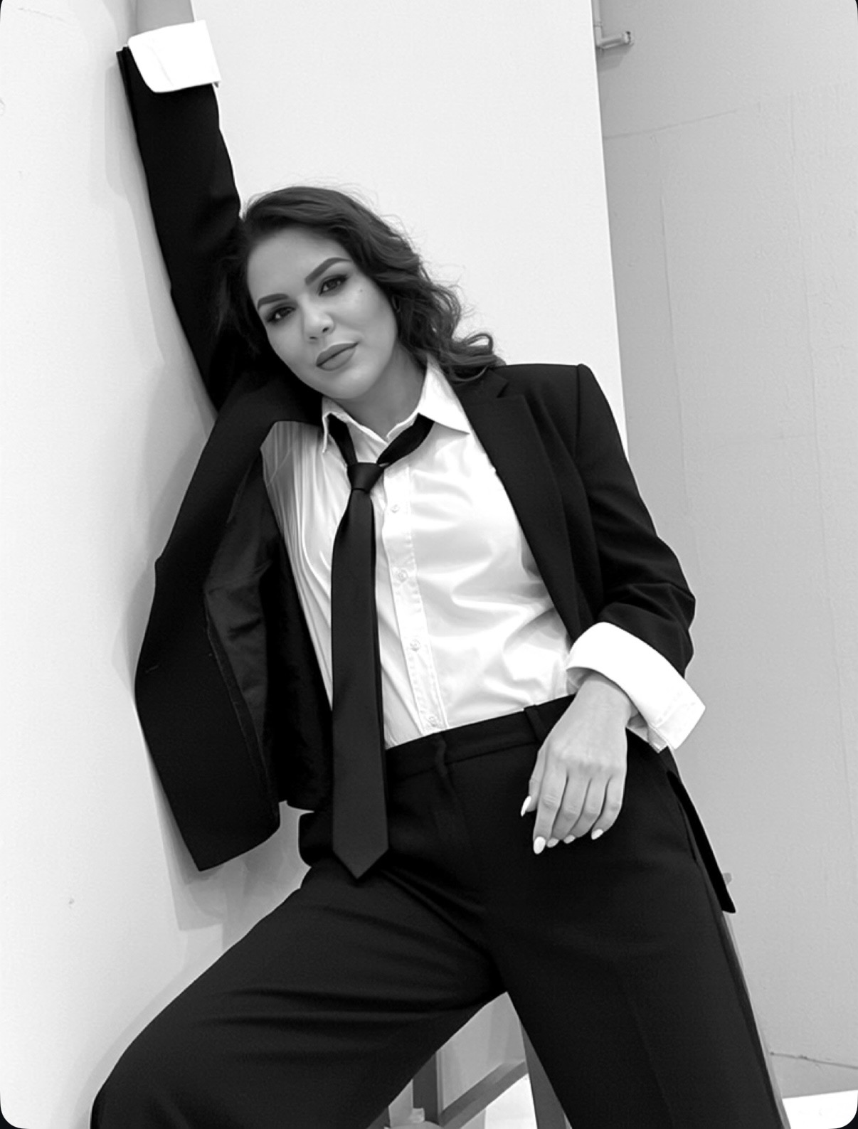 Woman in formal attire leaning against wall, one arm raised, looking at camera, black and white photo.