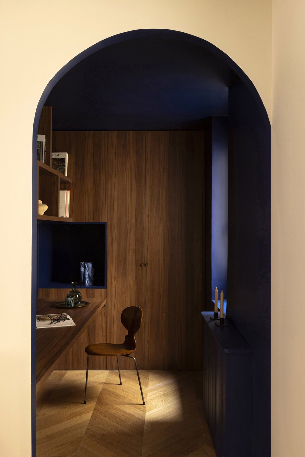 Interior view through an arched doorway showing a wooden door, a chair, a small desk, and shelves with books, illuminated by warm lighting.