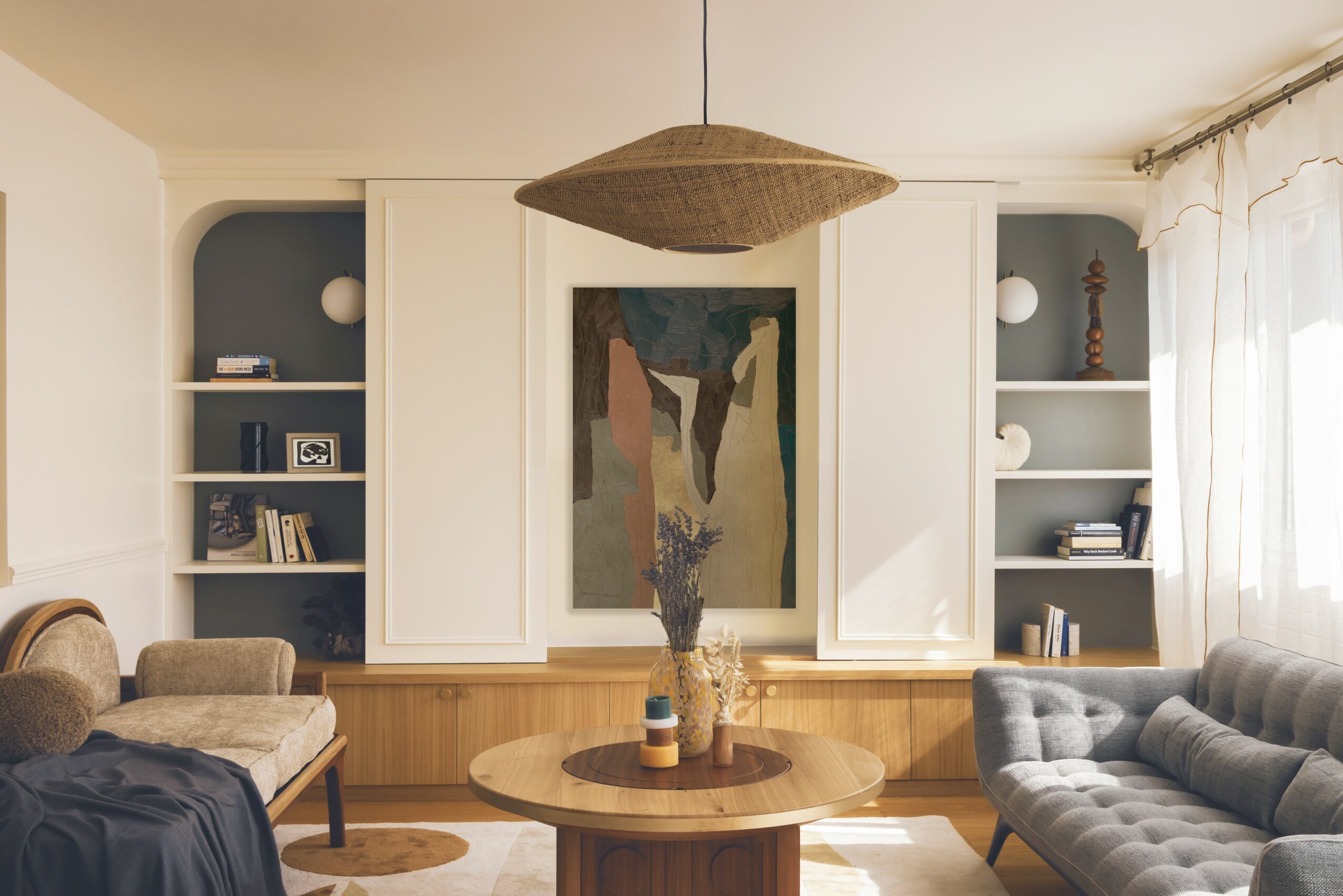 Living room with two sofas, a round wooden table, built-in shelves, and a large framed artwork, sunlight through curtains.