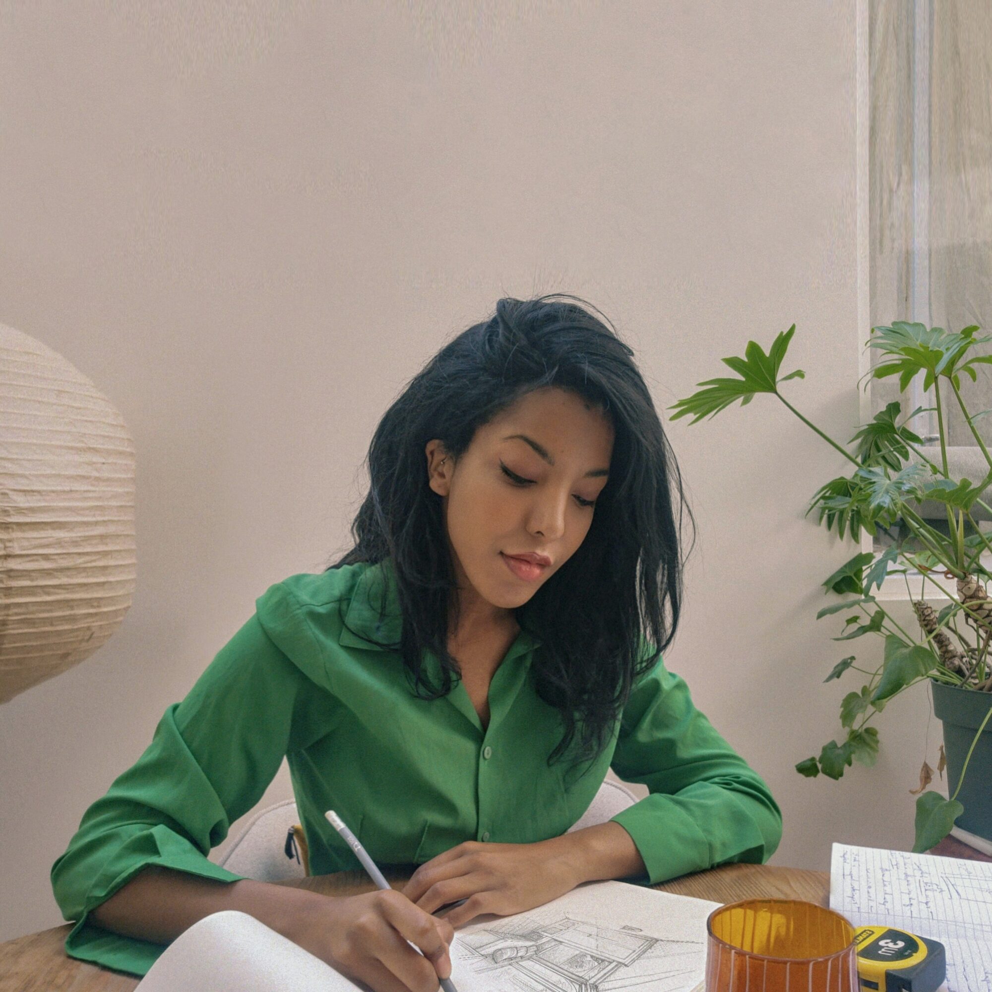 Woman with dark hair in a green shirt sitting at a table with a pen, paper, and a plant nearby.