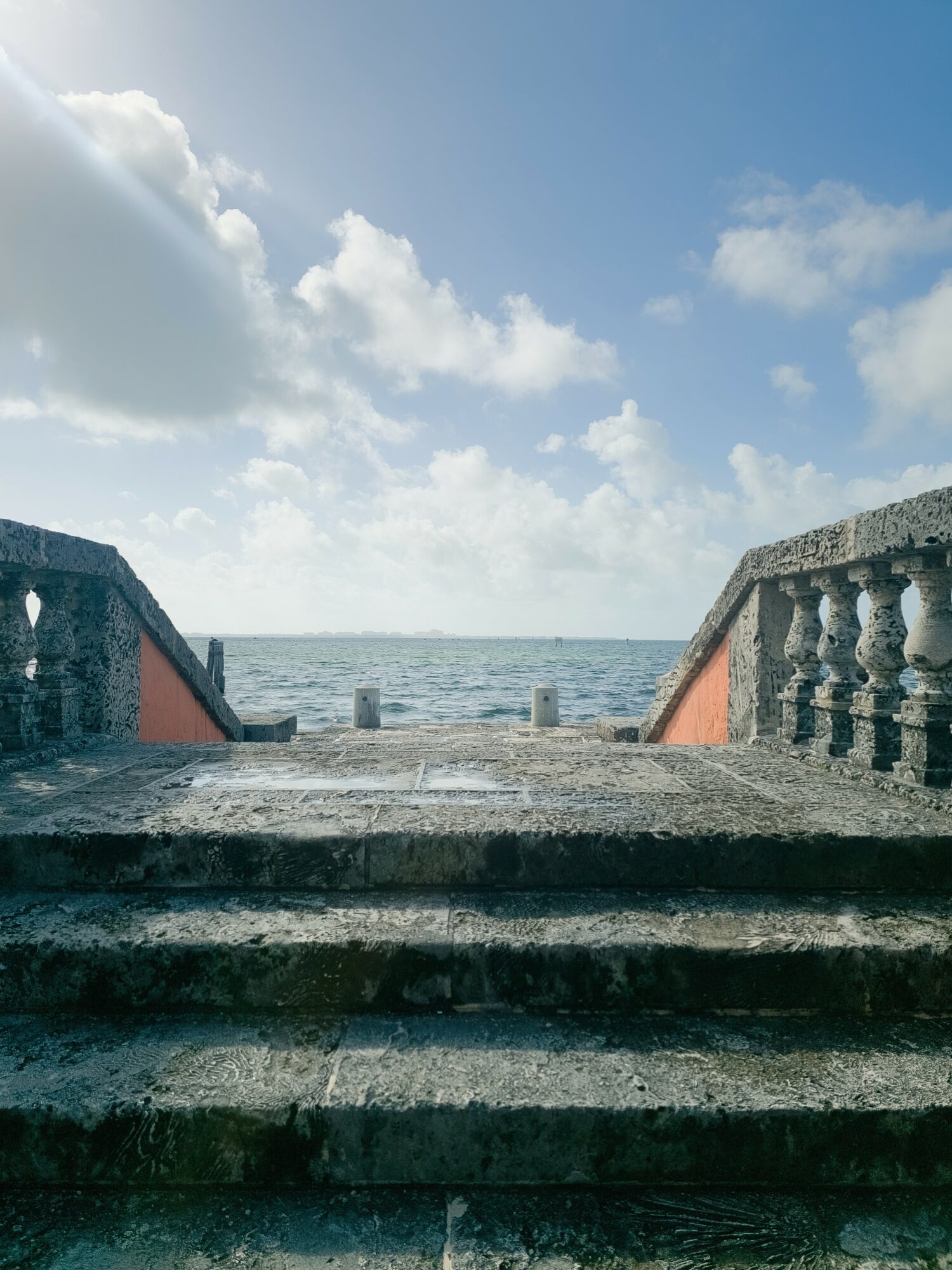 Stone steps leading to a view of the ocean with a partly cloudy sky, stone railings on sides, and a horizon line.