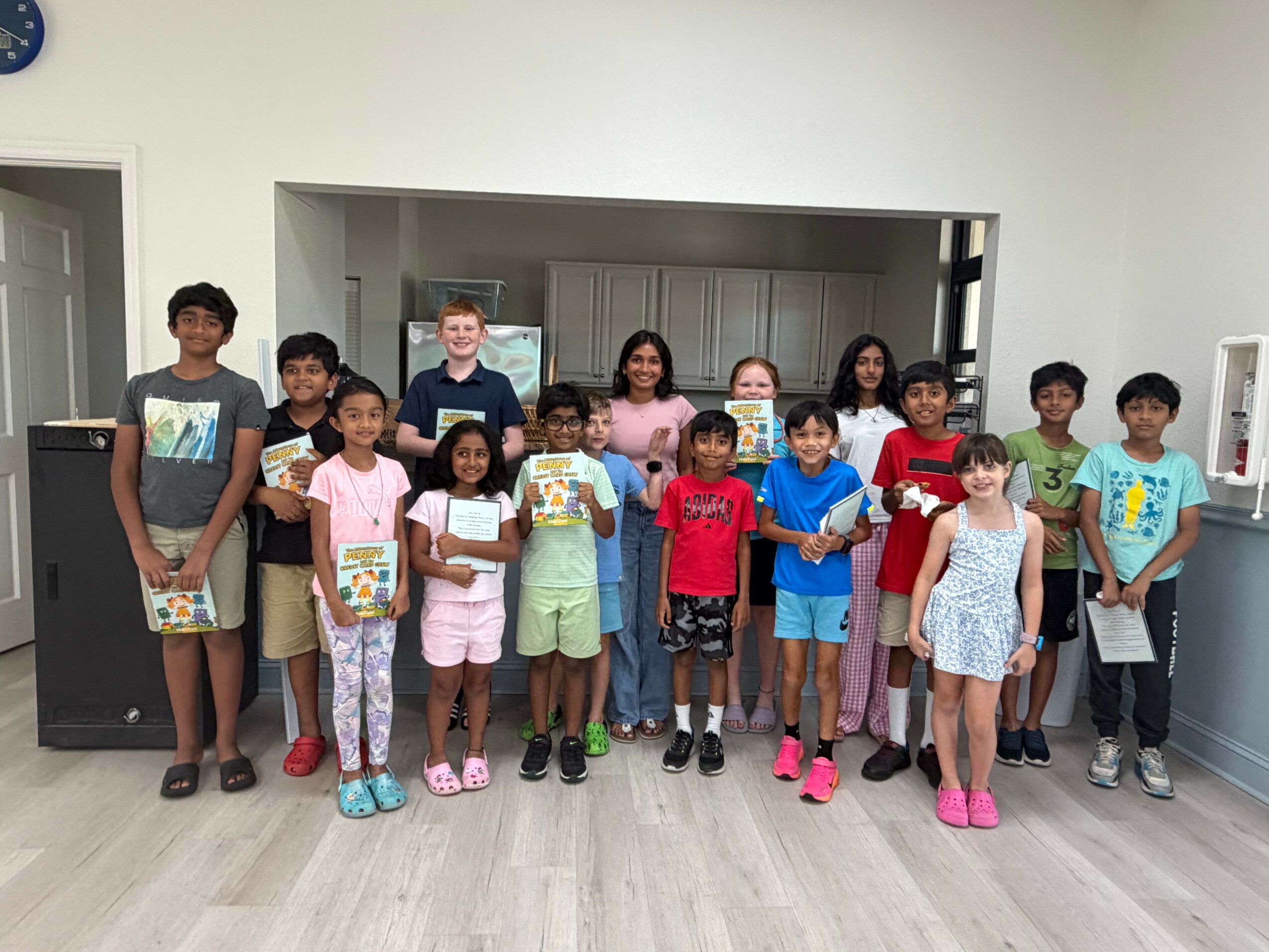 Group of children standing indoors, holding books or papers, smiling, in a room with white walls and wooden floor.