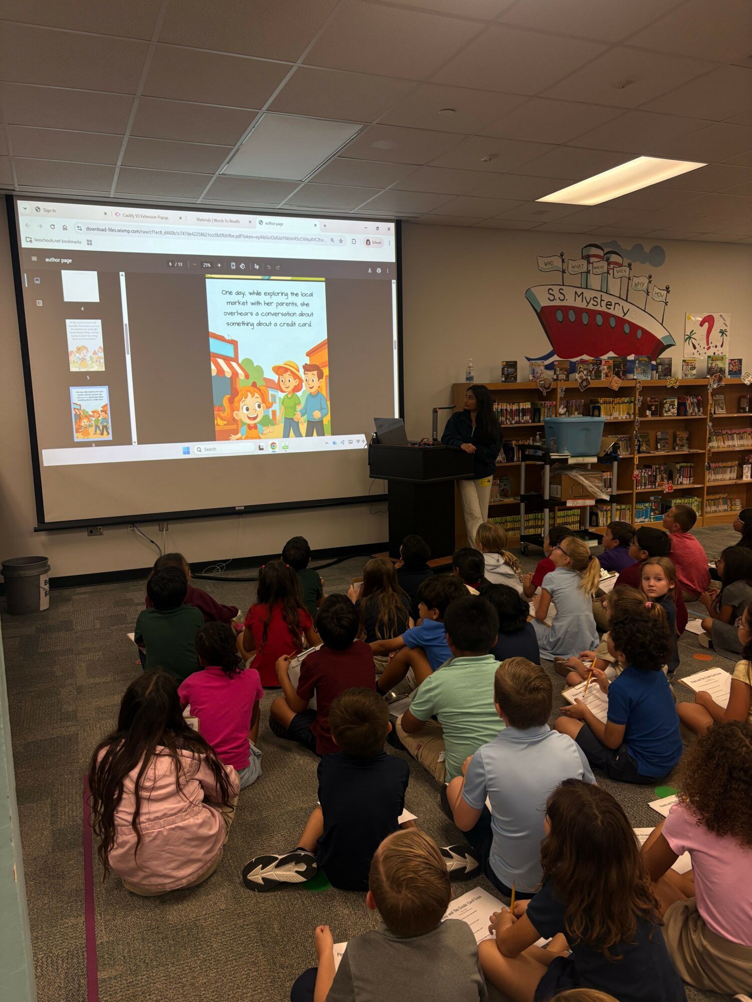 Group of children and adults gathered in a room watching a presentation on a screen, with bookshelves in the background.