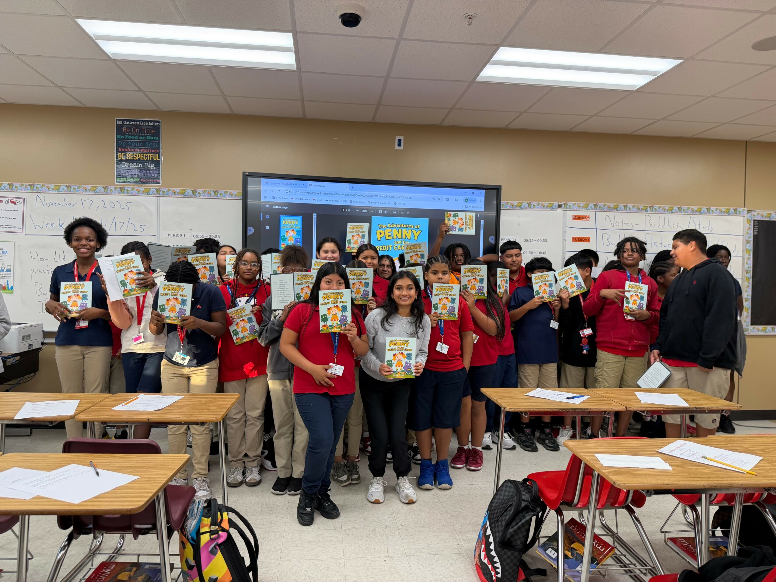 Group of students and teachers in a classroom holding books, with desks and a large screen behind them.