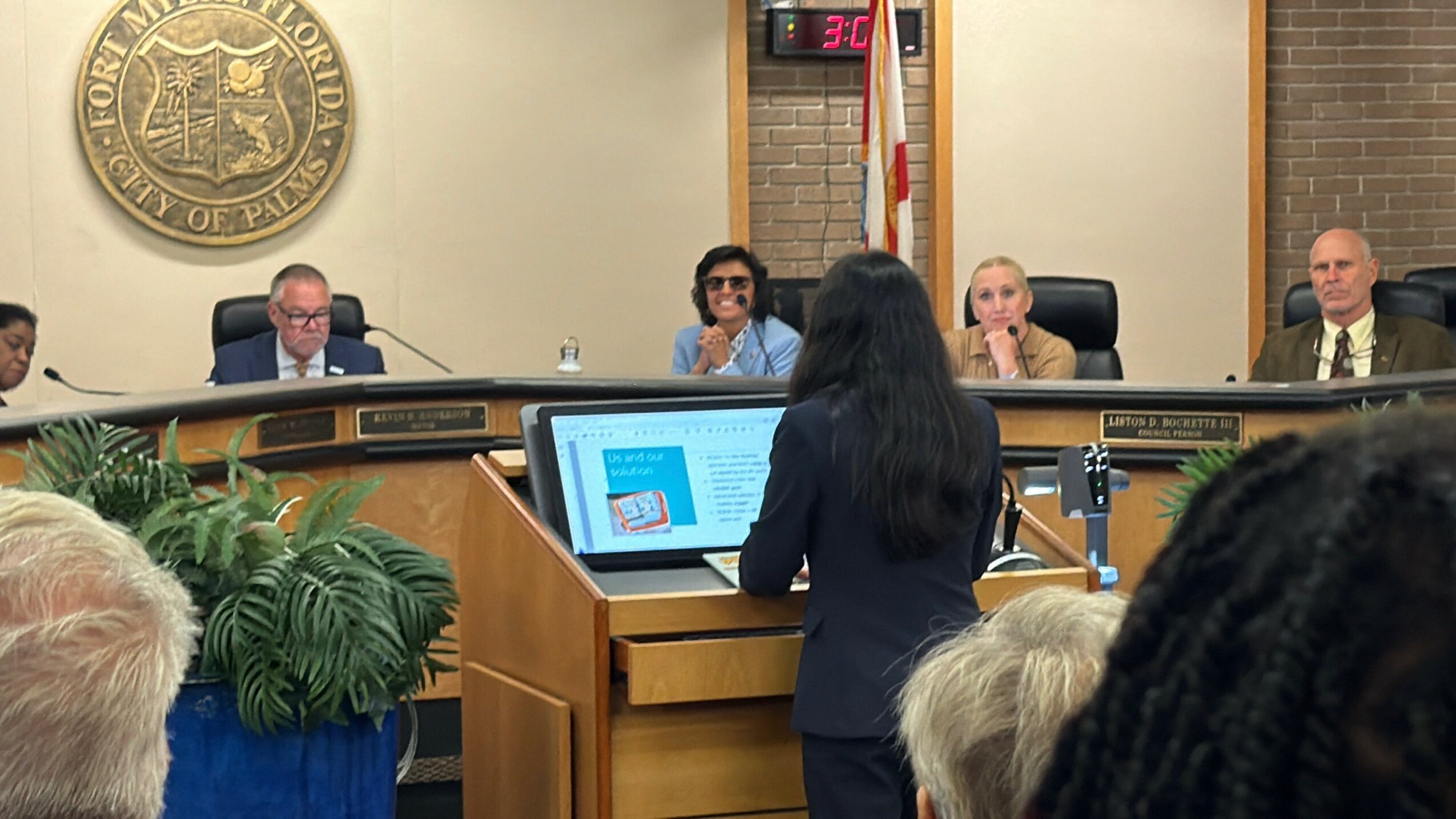 Person speaking at podium in a meeting room with five seated individuals behind, a large emblem on the wall, and a digital clock.