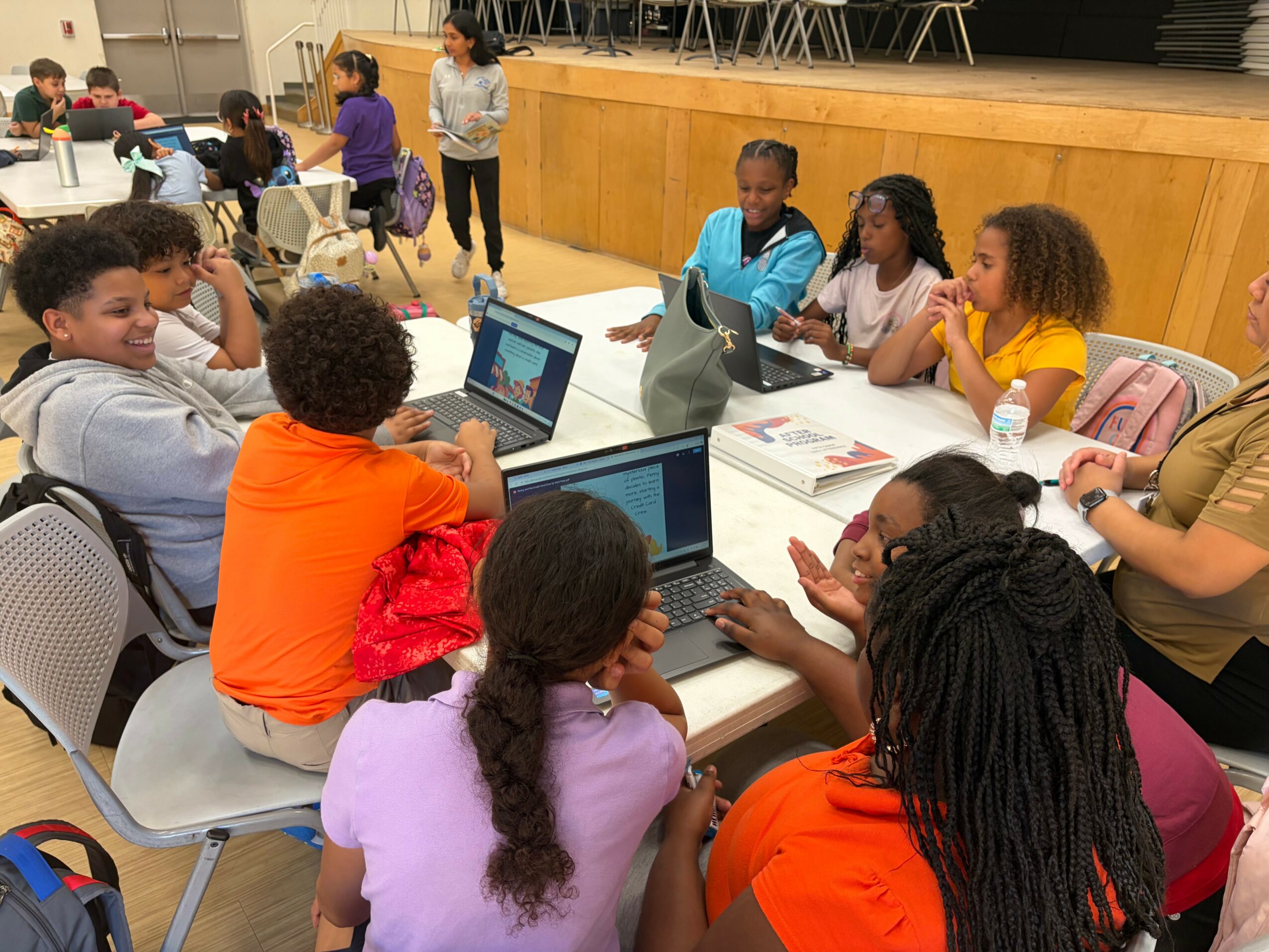 Group of children and teenagers sitting around a table with laptops, engaging in a discussion or activity, in a classroom or community space.