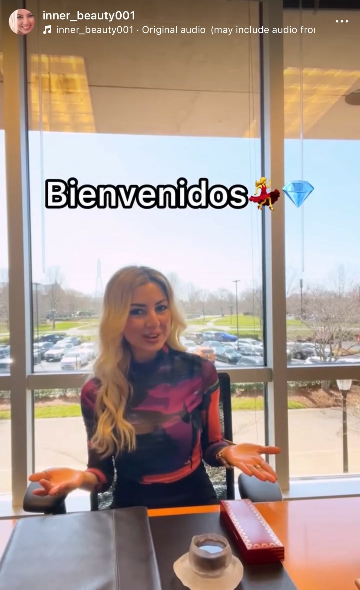 Woman with long blonde hair sitting at a table in front of large window, gesturing with hands, with a welcome message.