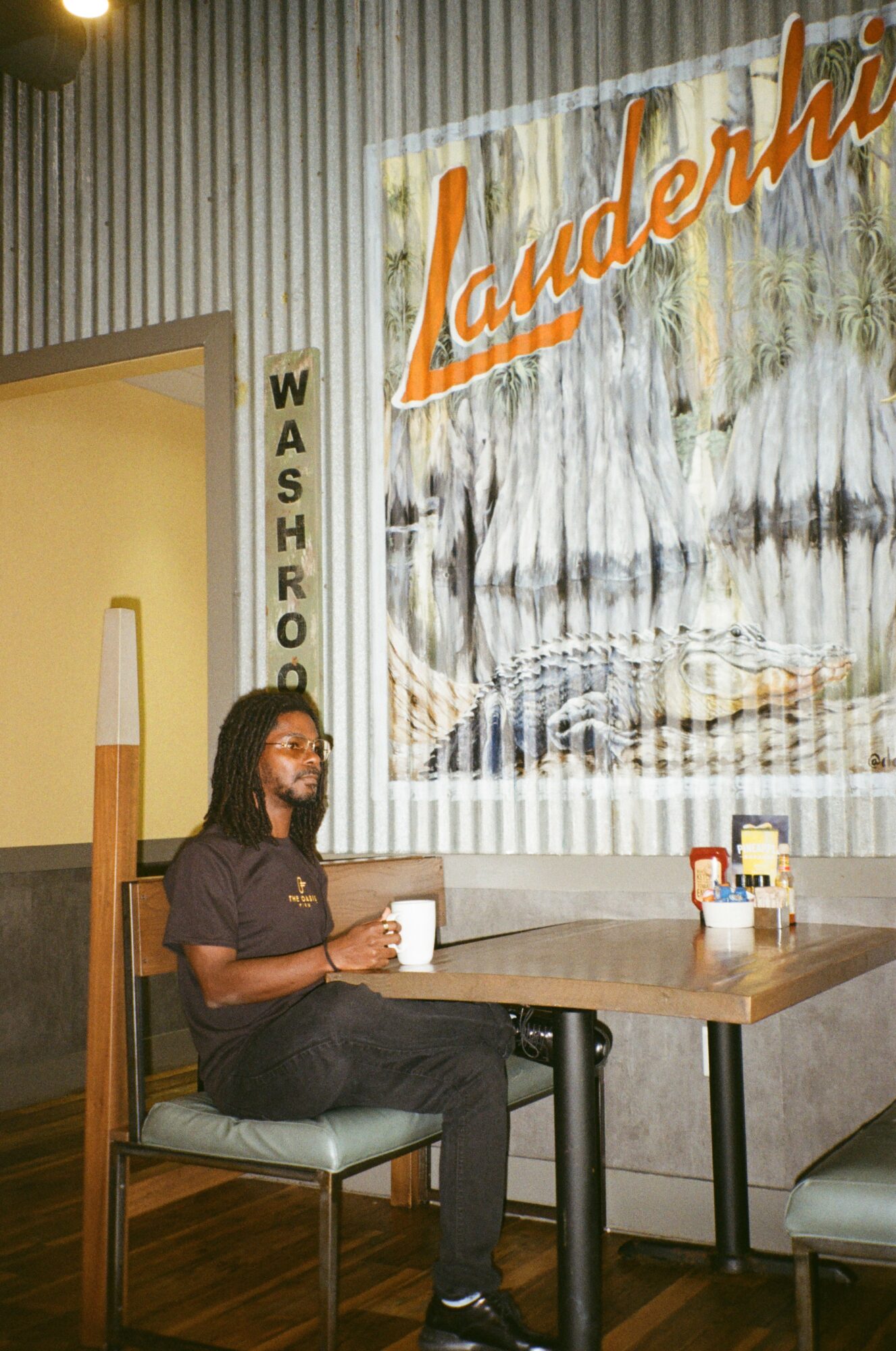 Woman sitting at a table in a laundromat, holding a cup, with a large sign reading 'Laundering' behind her.