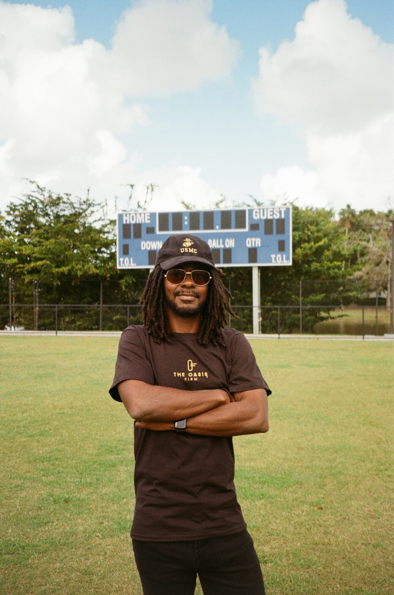 Person with long hair, sunglasses, and a hat standing on a grassy field with a scoreboard in the background.