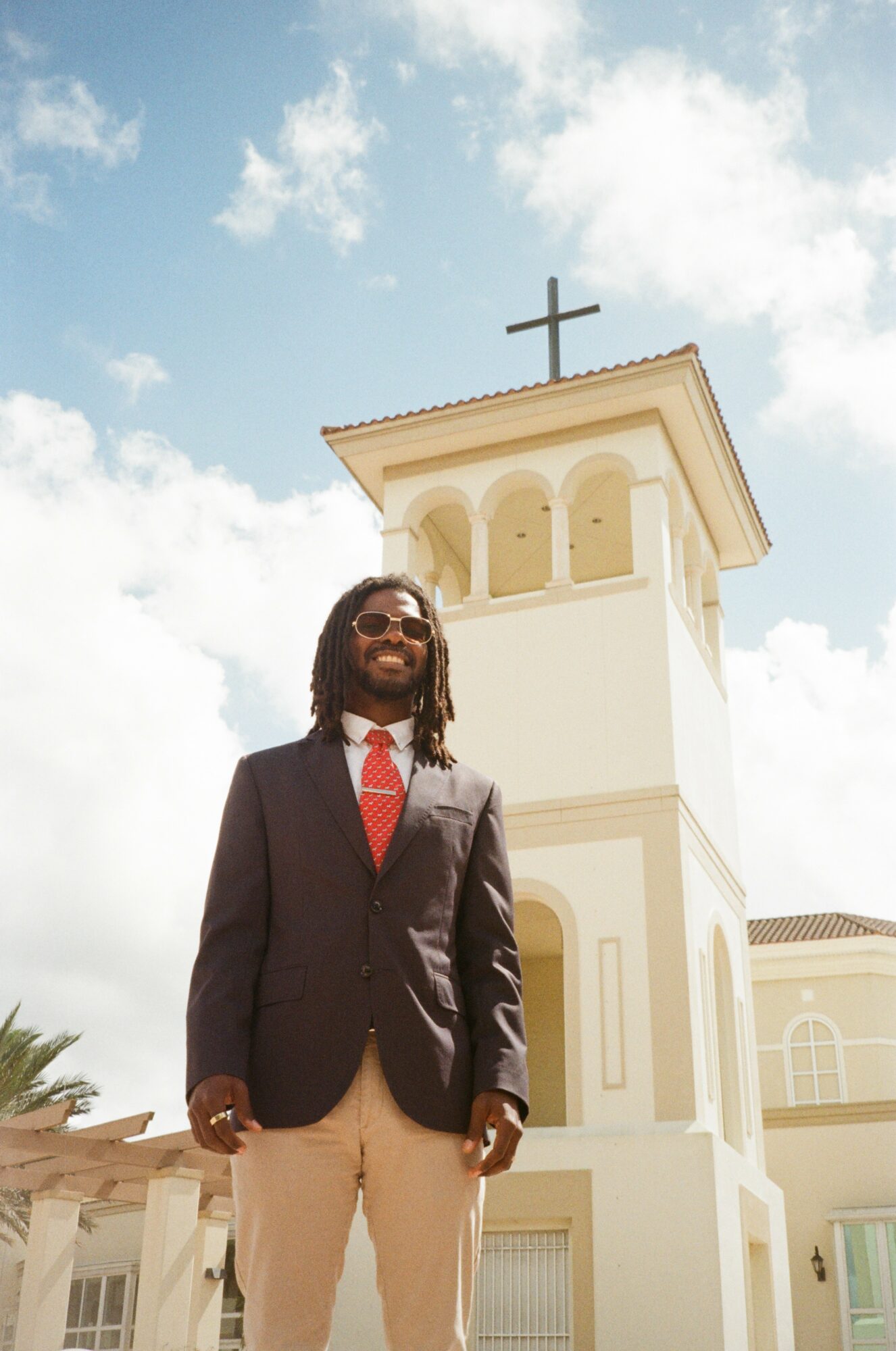 Man in suit and sunglasses standing outdoors in front of a white church with a cross on top, blue sky background.