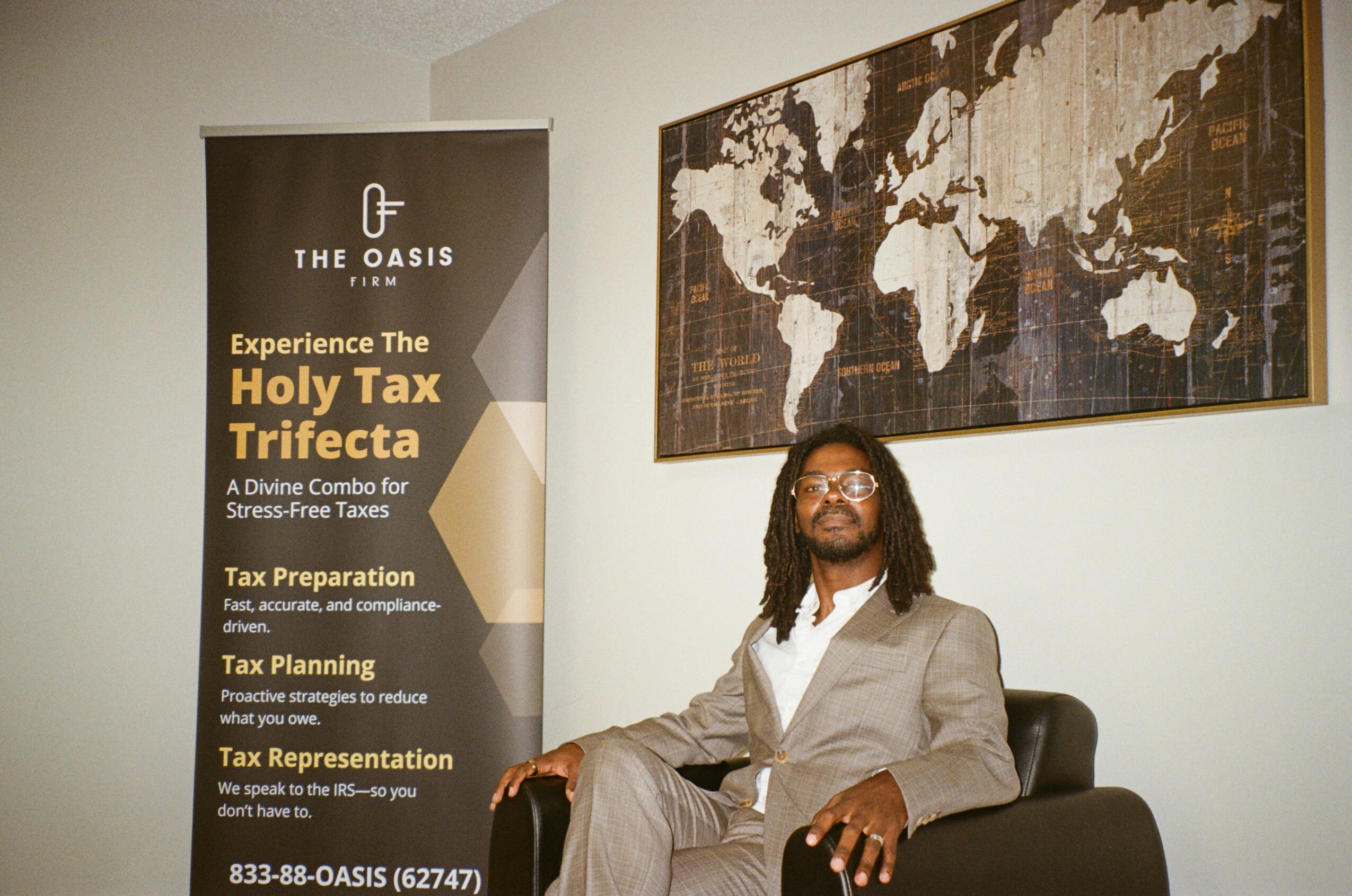 Man in a gray suit sitting on a black chair in an office, with a world map on the wall and a banner about taxes.