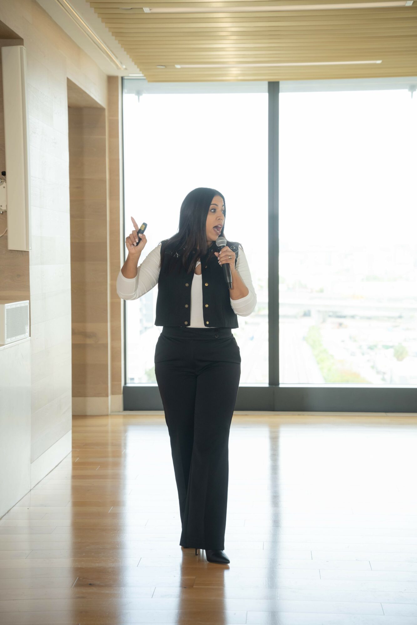 Woman standing indoors near large window, holding a microphone and pointing with her finger.