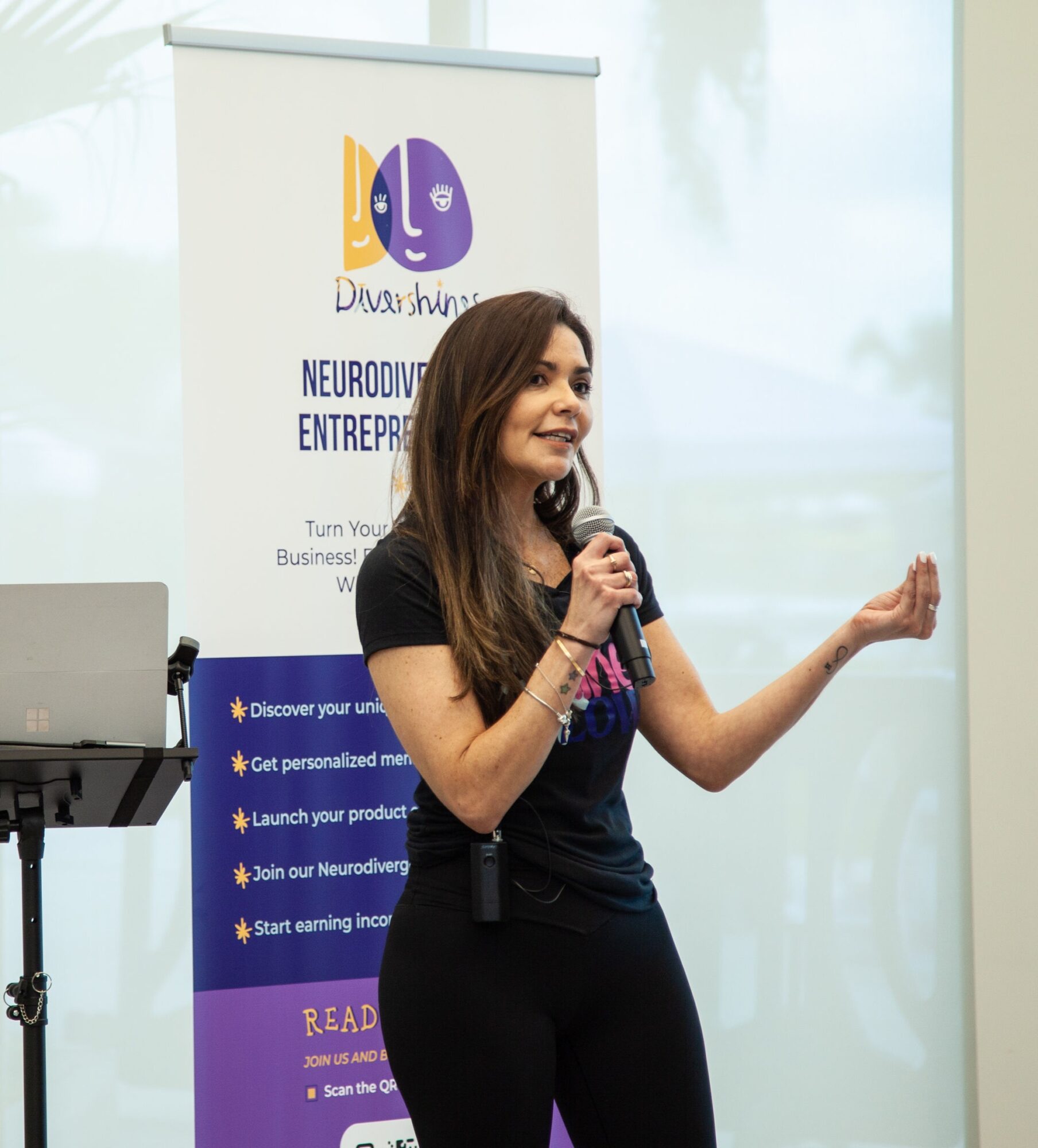Woman speaking into a microphone at an indoor event, with a colorful banner behind her.