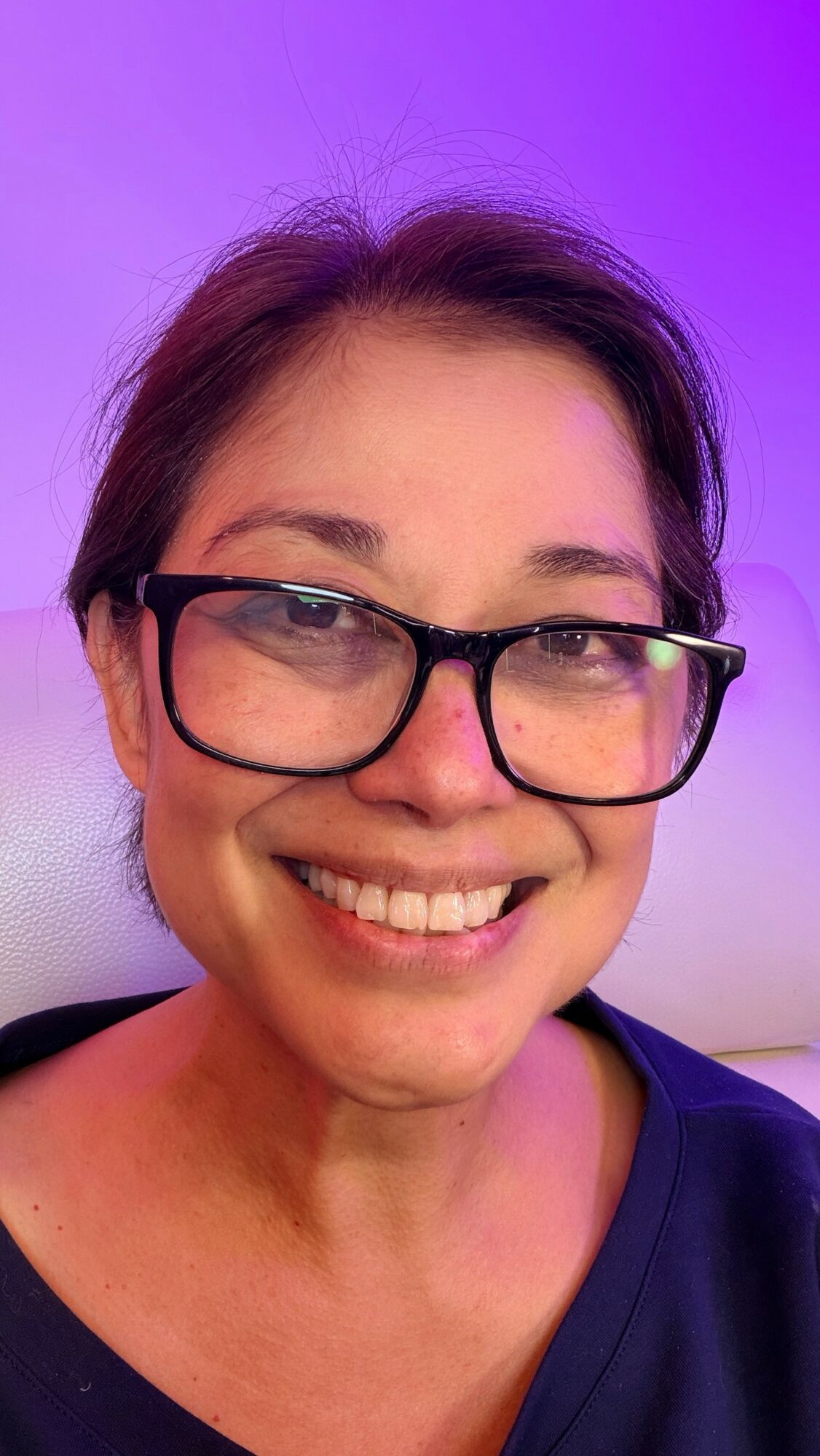 Smiling woman with glasses and short hair, purple background, wearing a dark top.