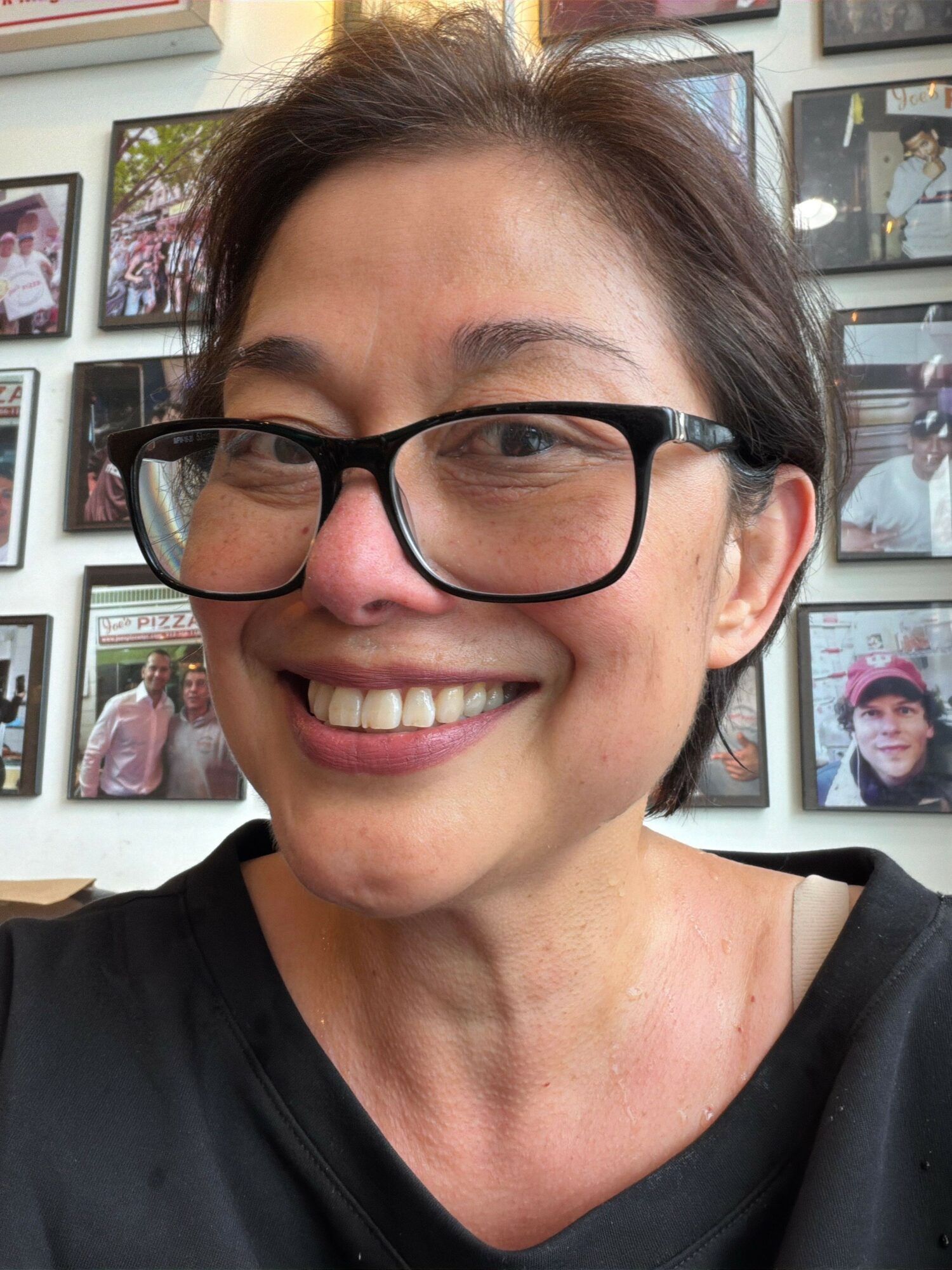 Smiling woman with glasses in front of a wall with framed photos, wearing a black top.