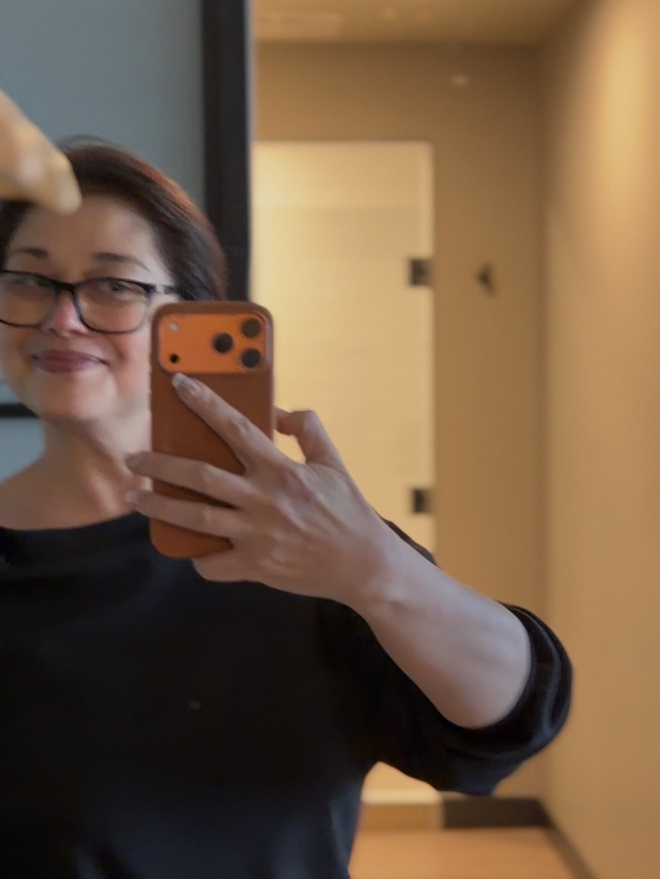 Person taking a mirror selfie with a smartphone, smiling, wearing glasses and a black top, in an indoor setting.
