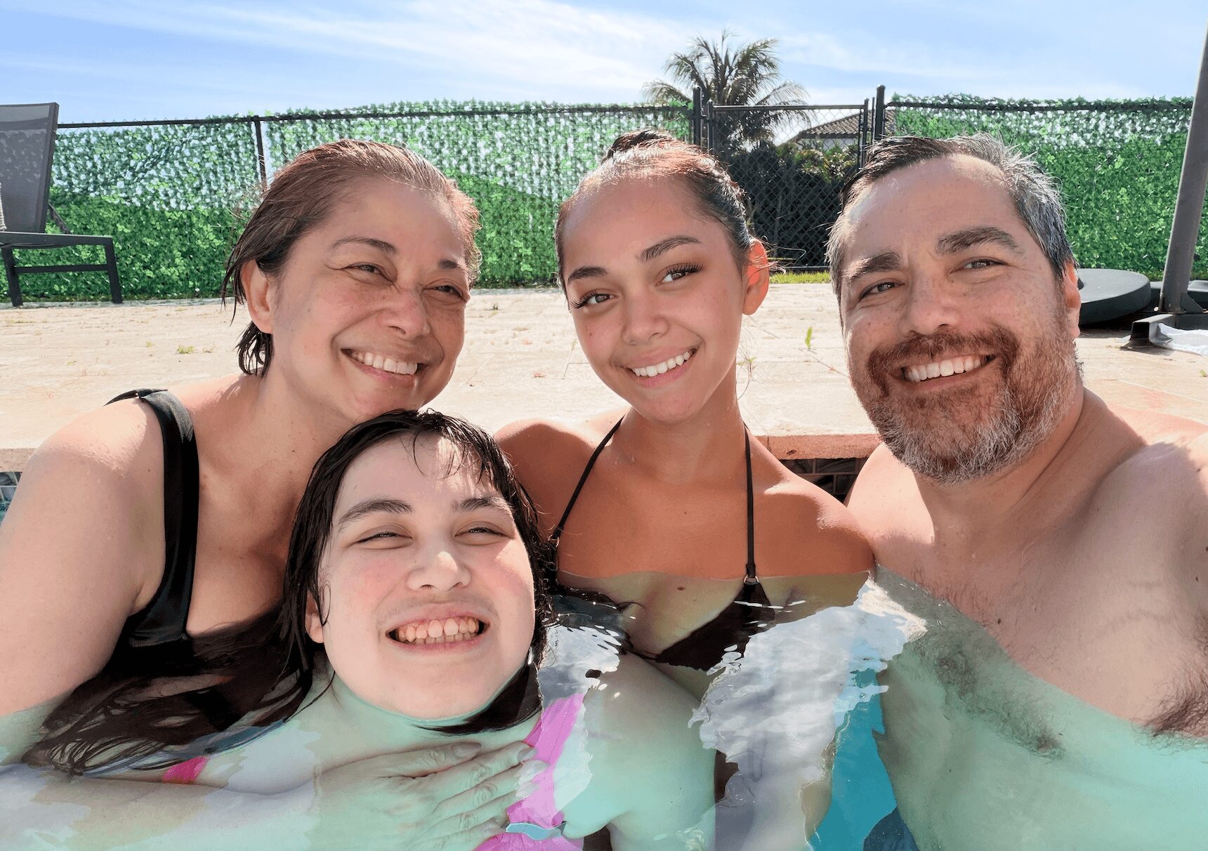 Four people smiling in a pool with a green fence and trees in the background.