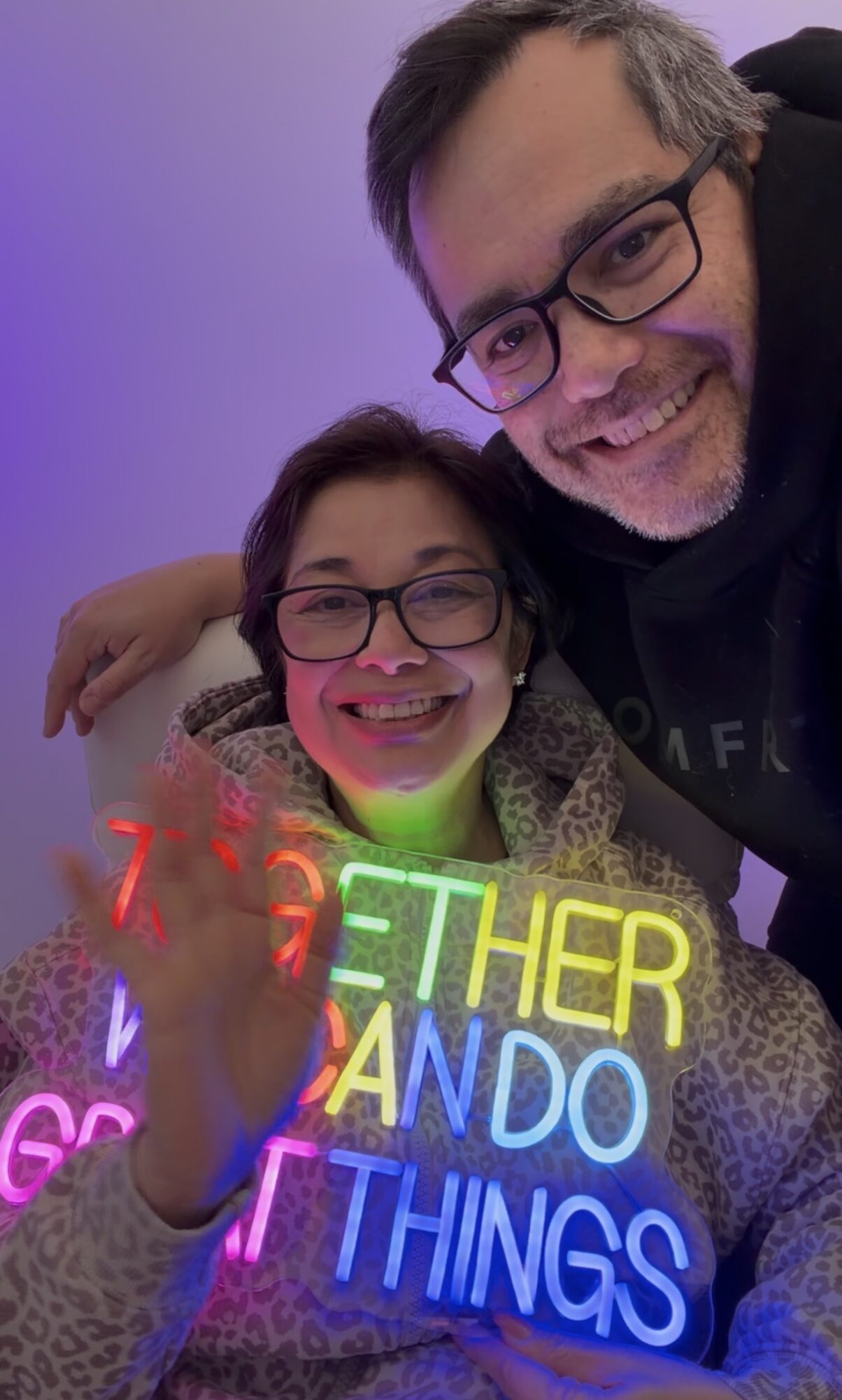 A man and a woman with glasses smiling, holding a colorful neon sign that reads 'Let her do anything,' against a purple background.