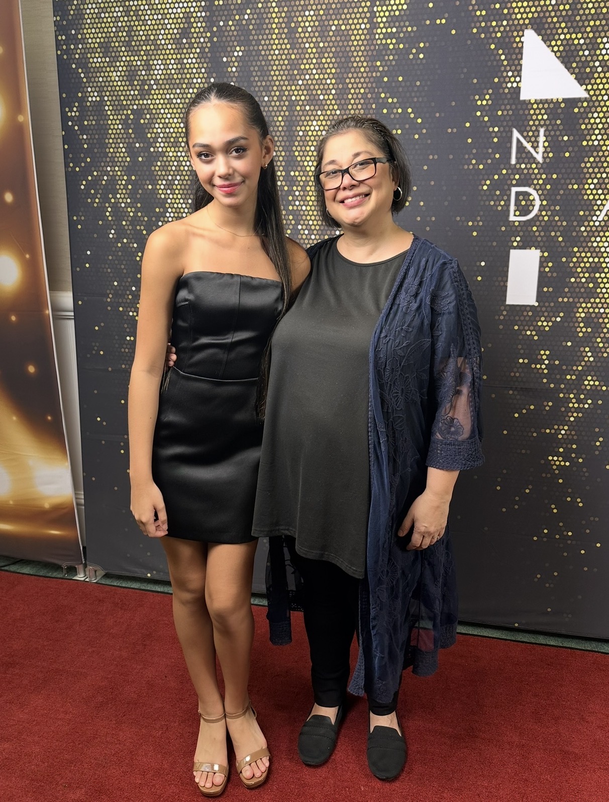 Two women standing on a red carpet in front of a black and gold backdrop, smiling at the camera.